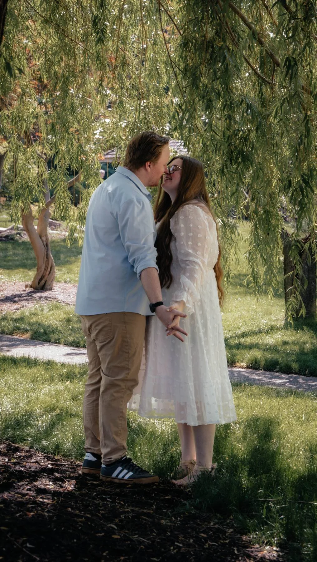A couple holding hands and leaning in to kiss under a leafy tree in a park.