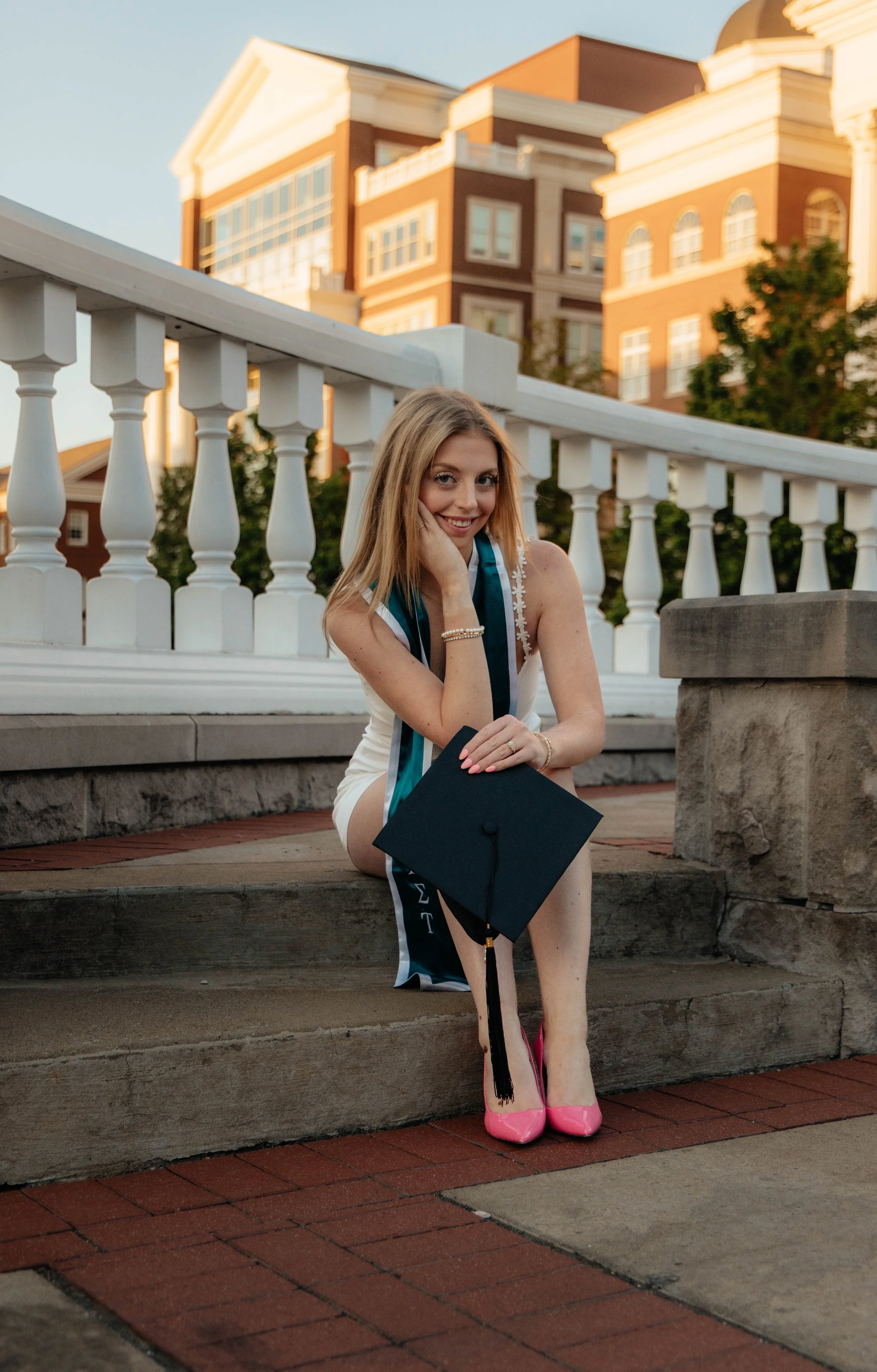 A young woman with blonde hair sitting on steps outdoors during sunset, wearing a graduation stole, holding her graduation cap, and smiling at the camera. She is wearing pink high heels and has a building with large windows and white architectural details in the background.