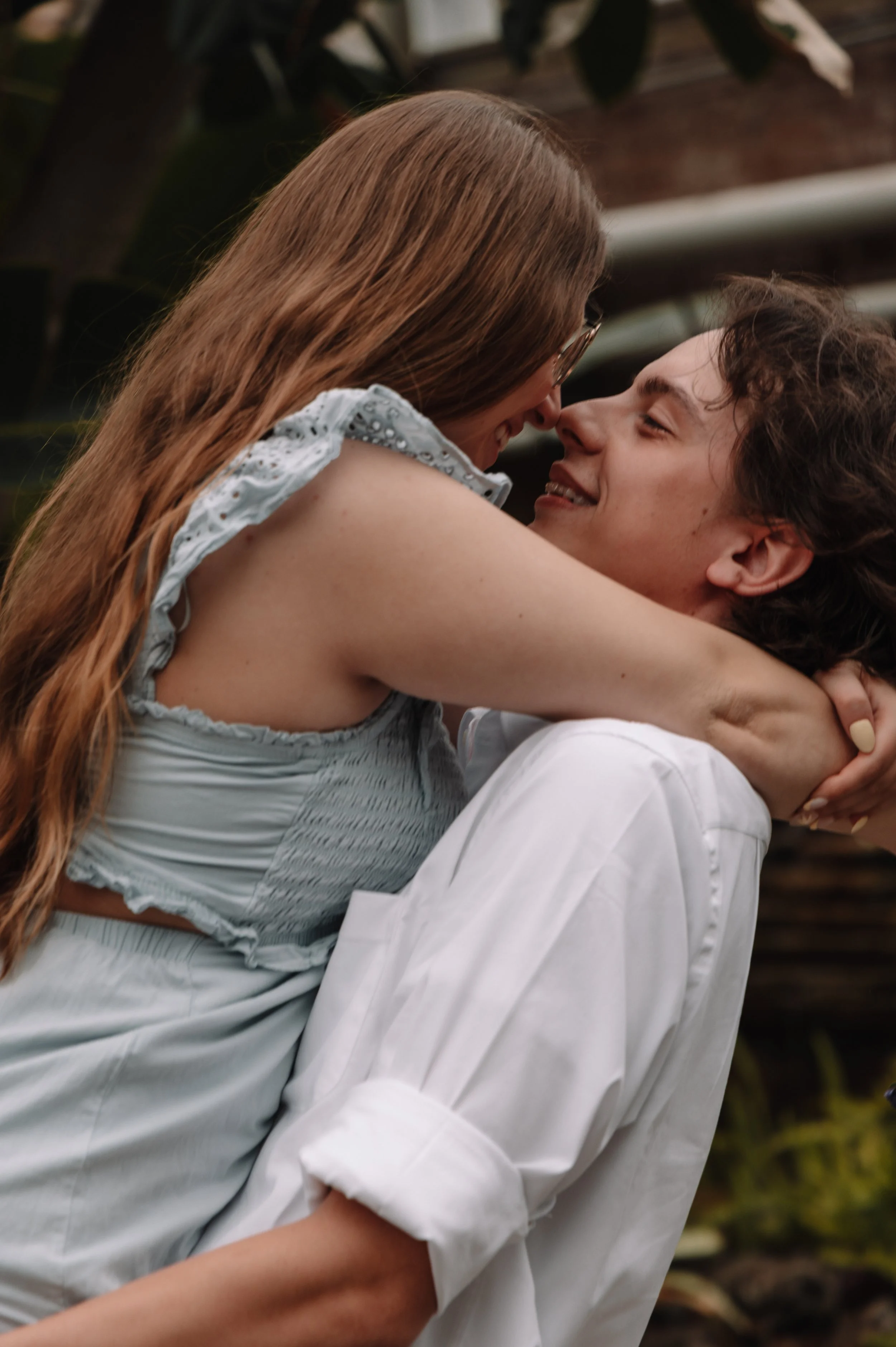 A couple with their faces close, smiling and embracing outdoors, with a blurred background of foliage.