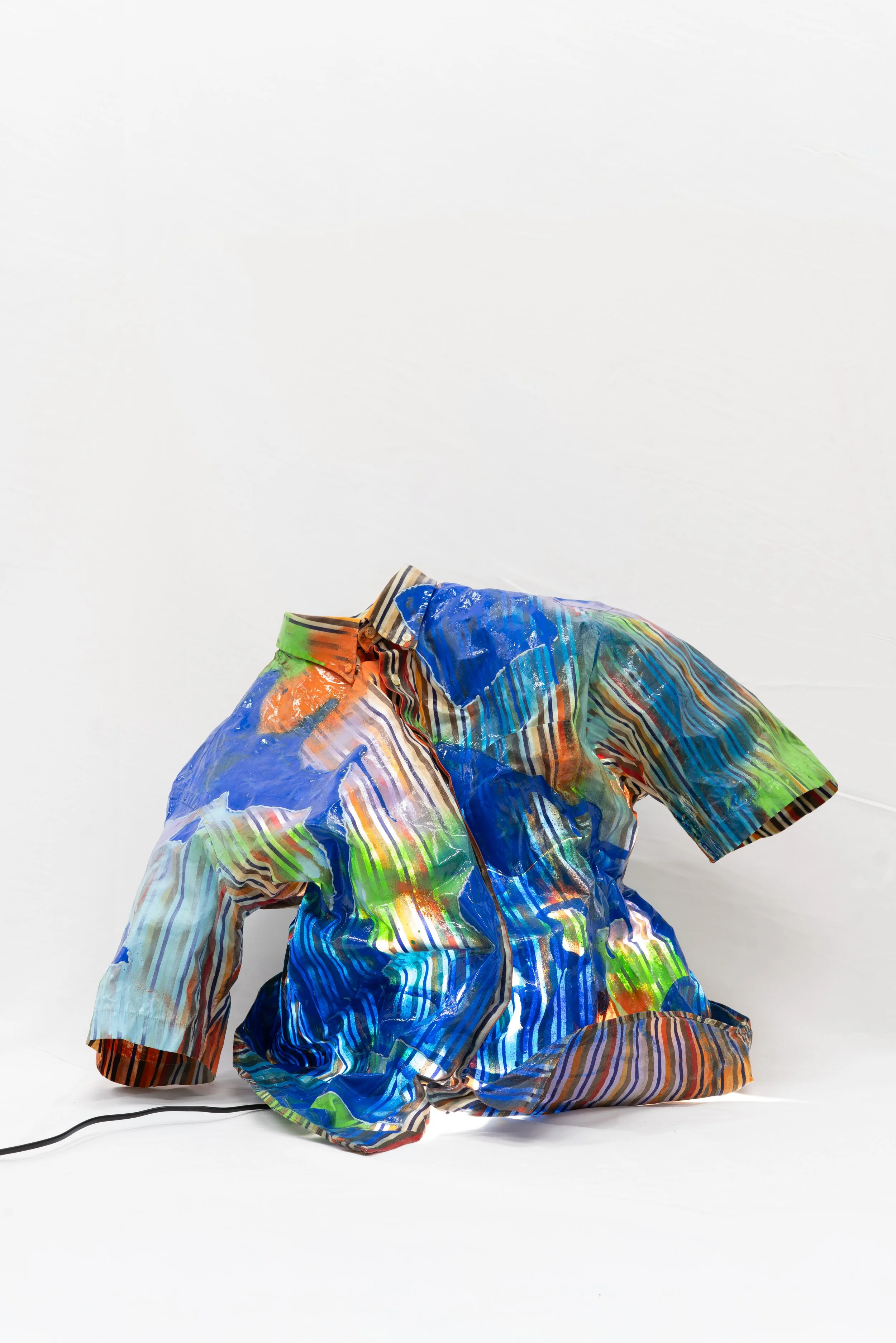 A person kneeling on the floor wearing a colorful, abstract patterned shirt. The shirt features various colors including blue, green, orange, and brown with a striped collar. The person's face is not visible, and the background is plain white.