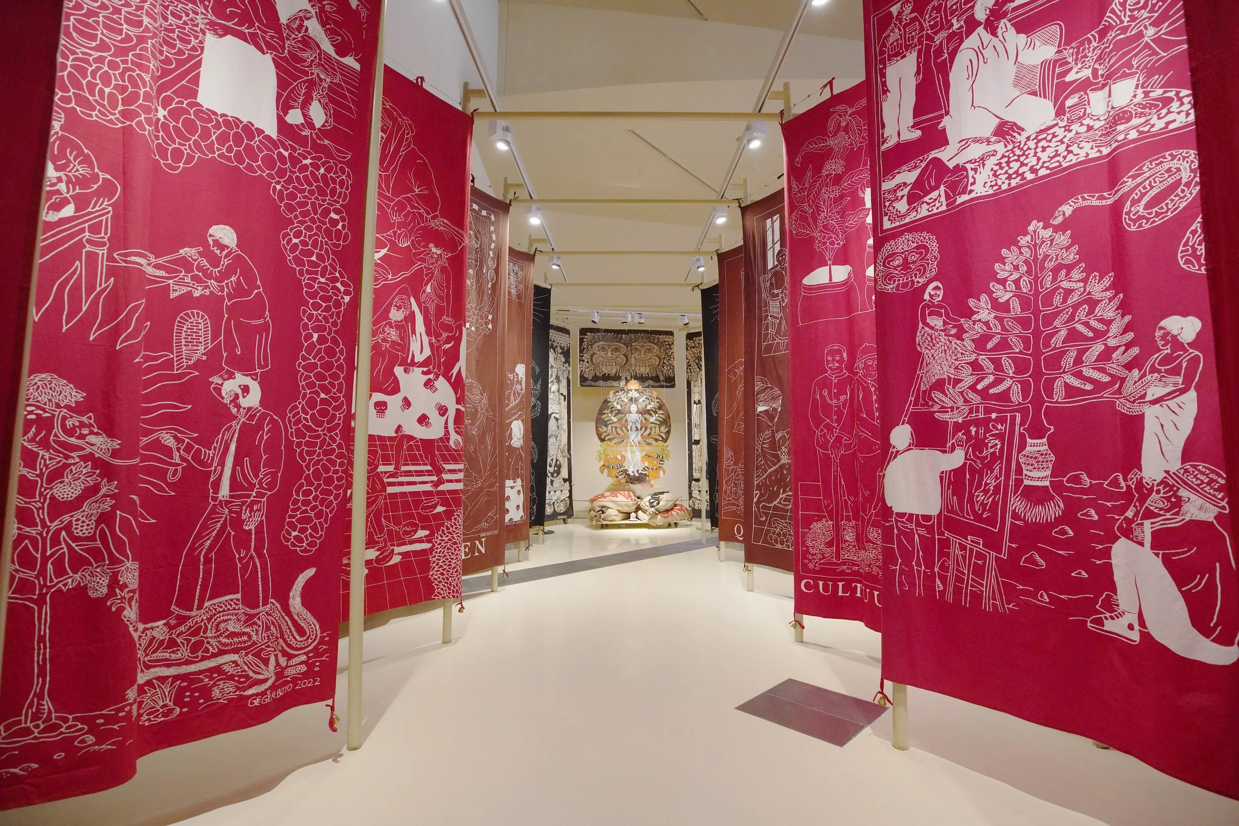 Exhibition with tall fabric panels featuring white line art illustrations on red backgrounds, displayed in a gallery with overhead lighting and a centerpiece artwork at the back.