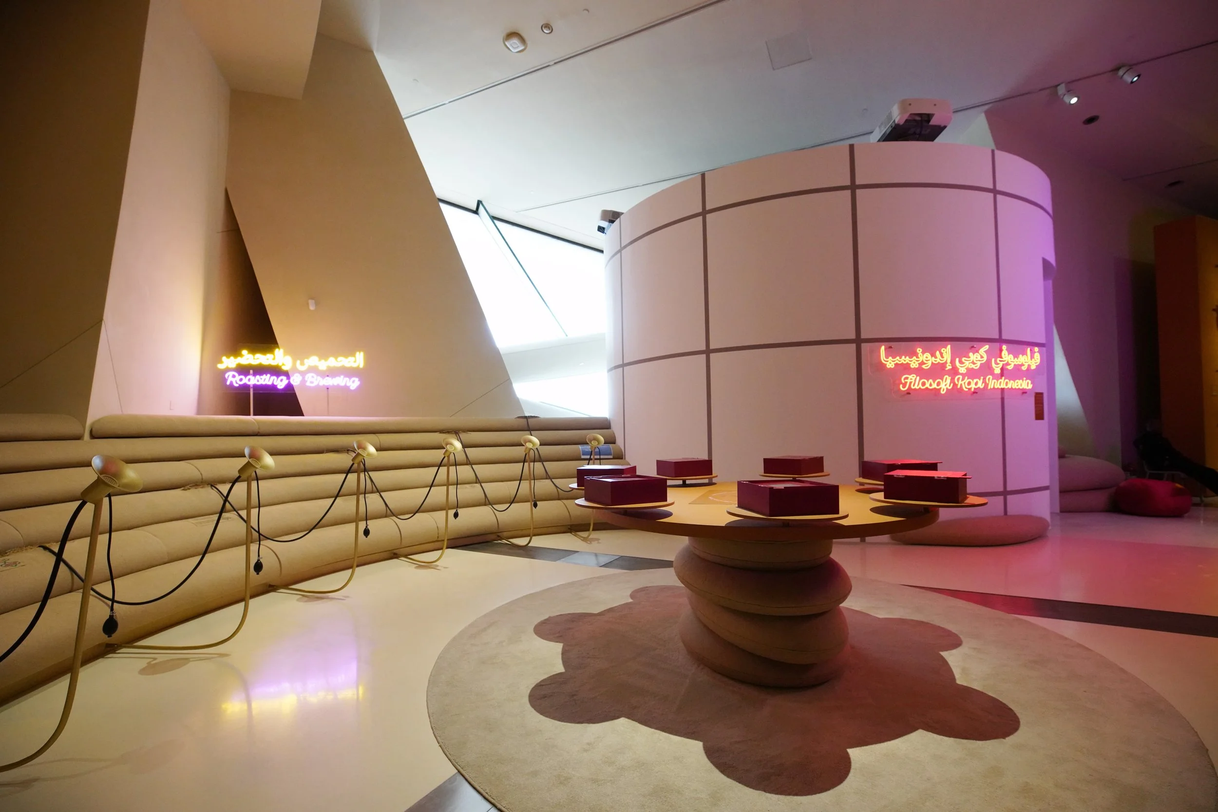 Interior of a modern space with beige curved seating, a round table with books, and colorful neon signs in Arabic and English. The signs say 'Roasting & Brewing' and 'Filoneg Kopi Indonesia.'