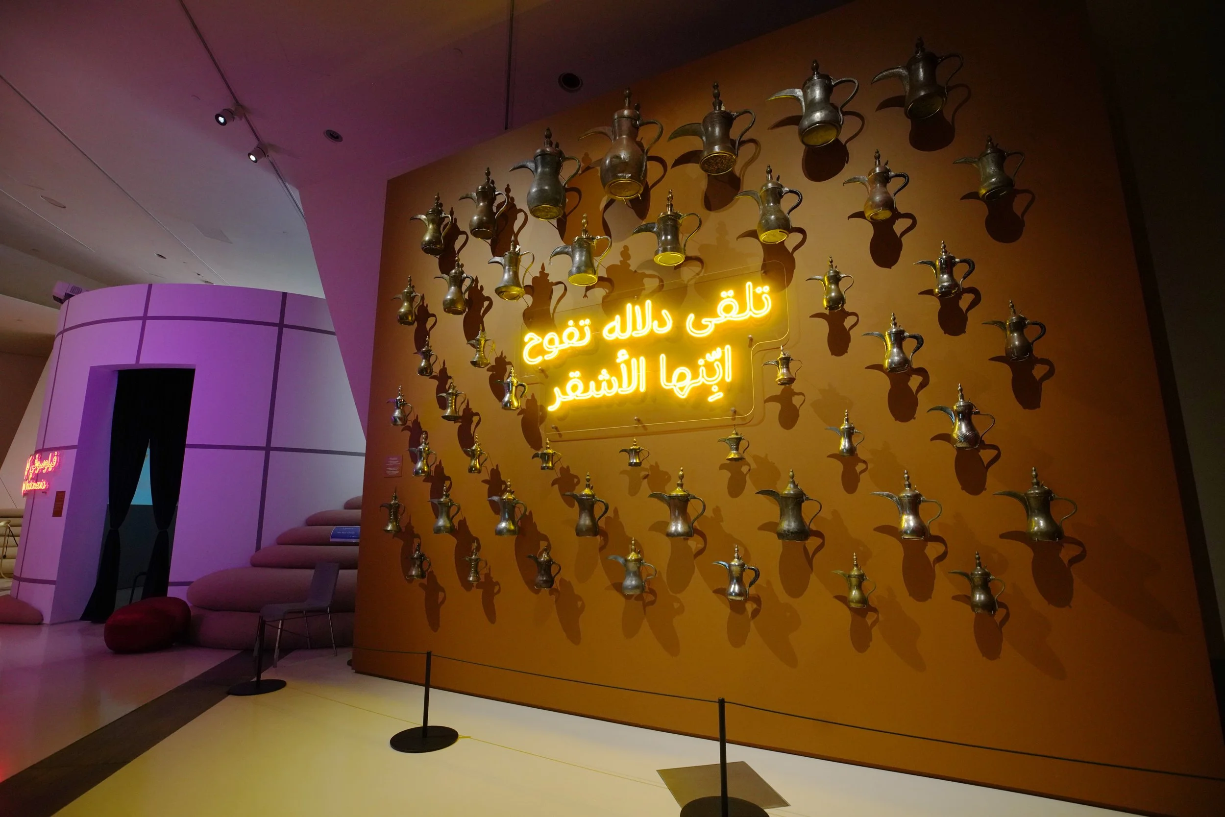 Wall display with numerous gold and silver traditional Middle Eastern teapots arranged around a yellow neon sign with Arabic text in a modern interior space.