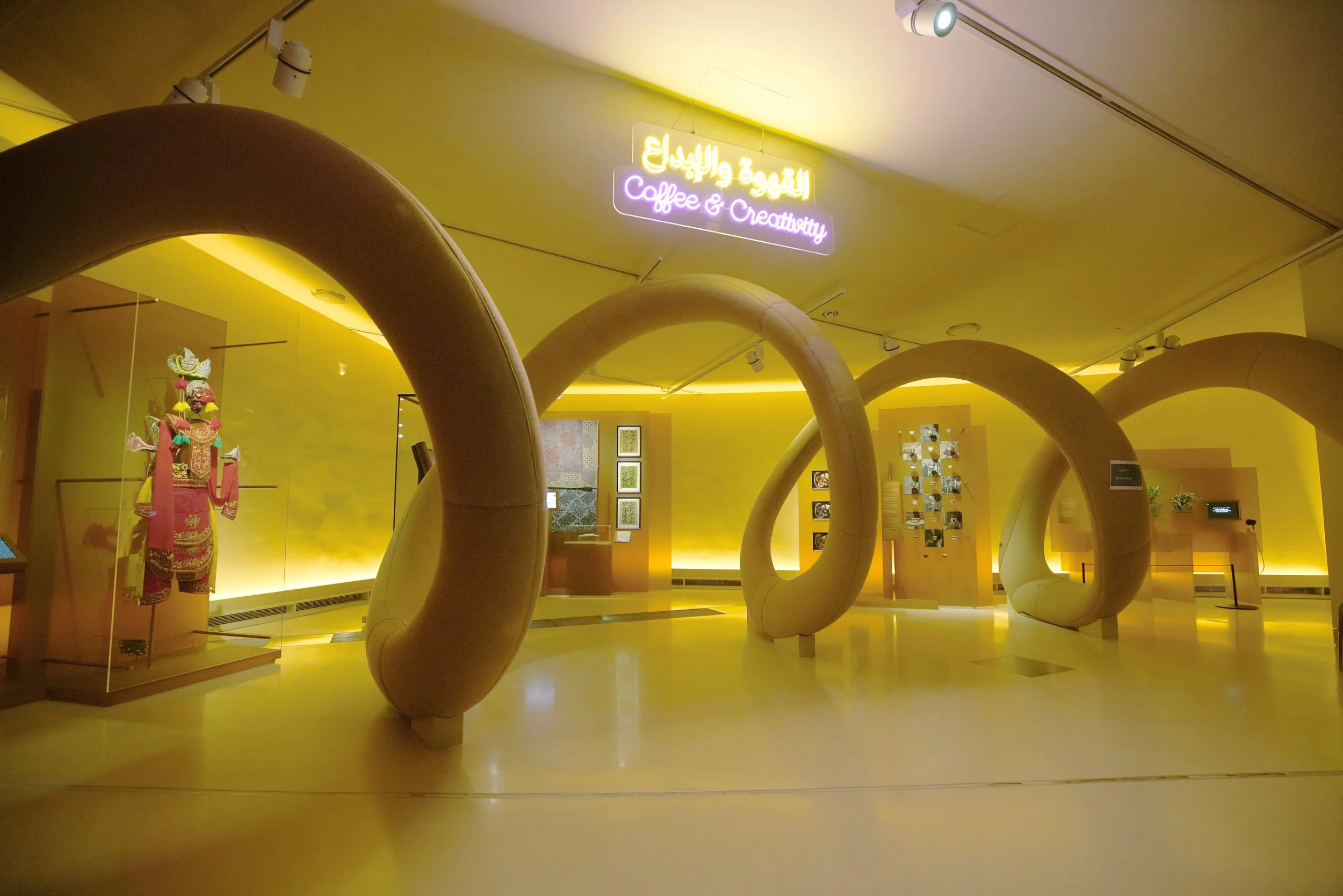 An exhibit with large, light-colored, spiral sculptures in a well-lit room with yellow walls. There is a neon sign on the ceiling that reads 'Coffee & Creativity' in English and Arabic. Displays with artwork and a traditional costume are visible in the background.