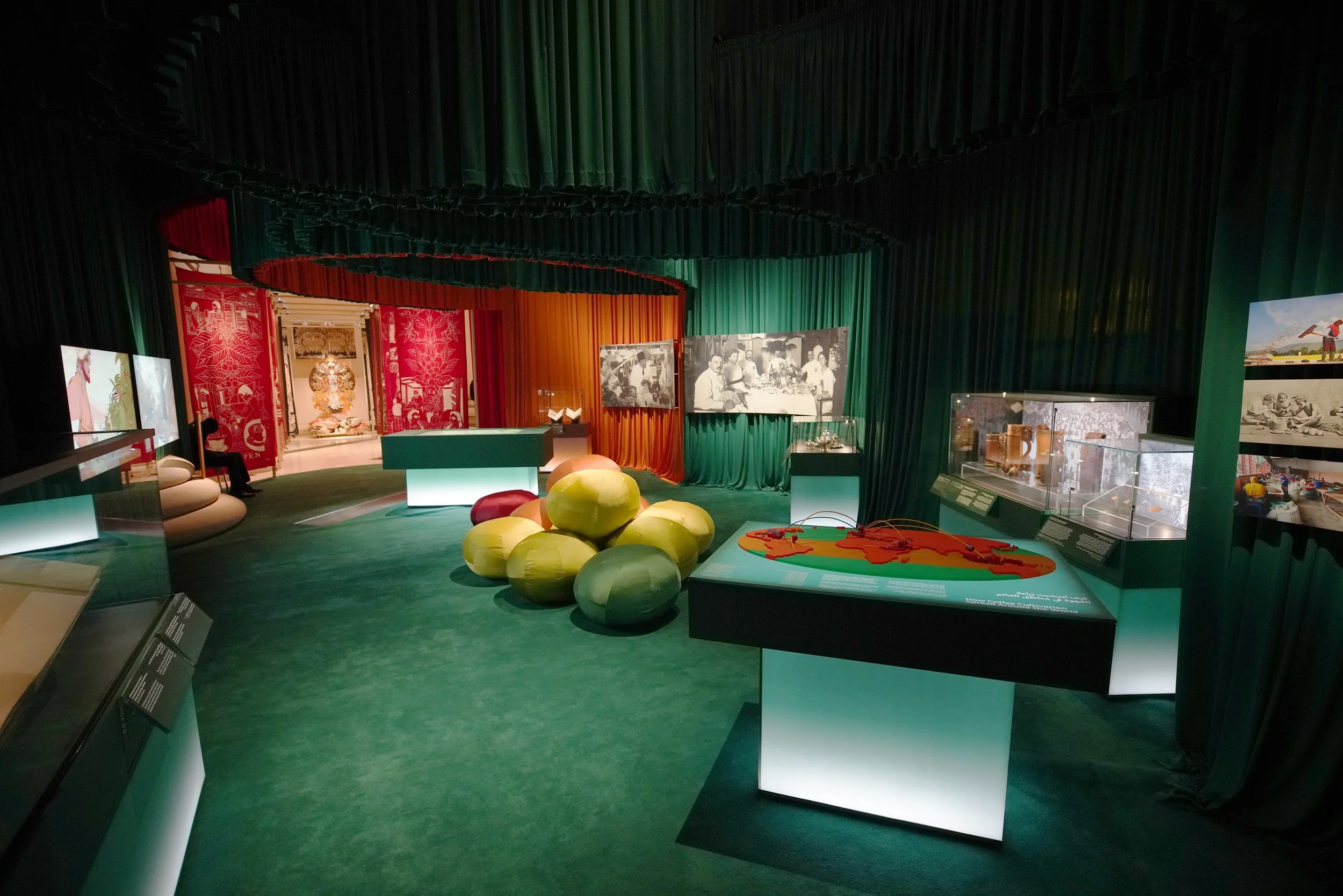 An exhibit room with green, red, and orange curtains, displays of historical artifacts and photographs, and a large green and yellow arrangement resembling a cluster of large spheres.