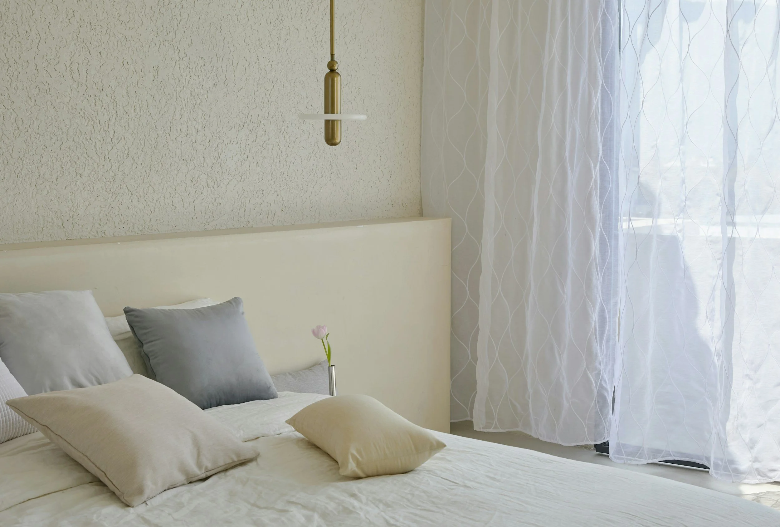 A bedroom with a bed, multiple pillows, a white headboard, a hanging light, a small vase with a pink tulip, and sheer white curtains next to a window.