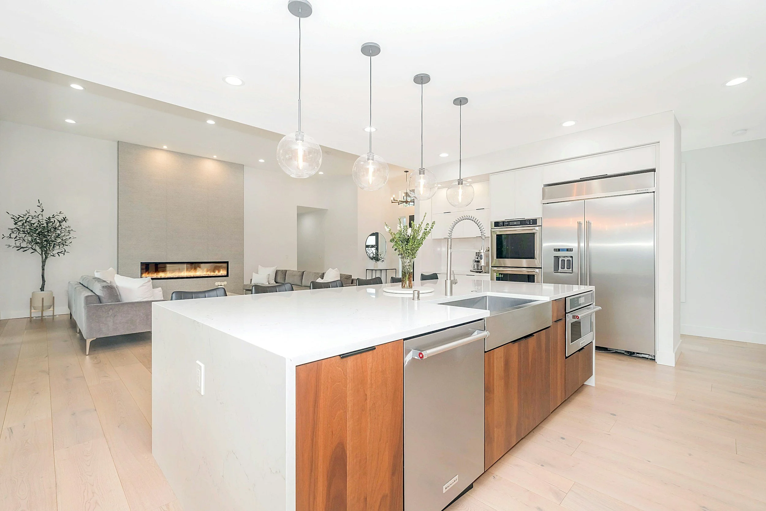 Modern open-concept kitchen with white island, stainless steel appliances, and pendant lights, connected to a living room with a hanging fireplace, gray sofas, and a tree decoration.
