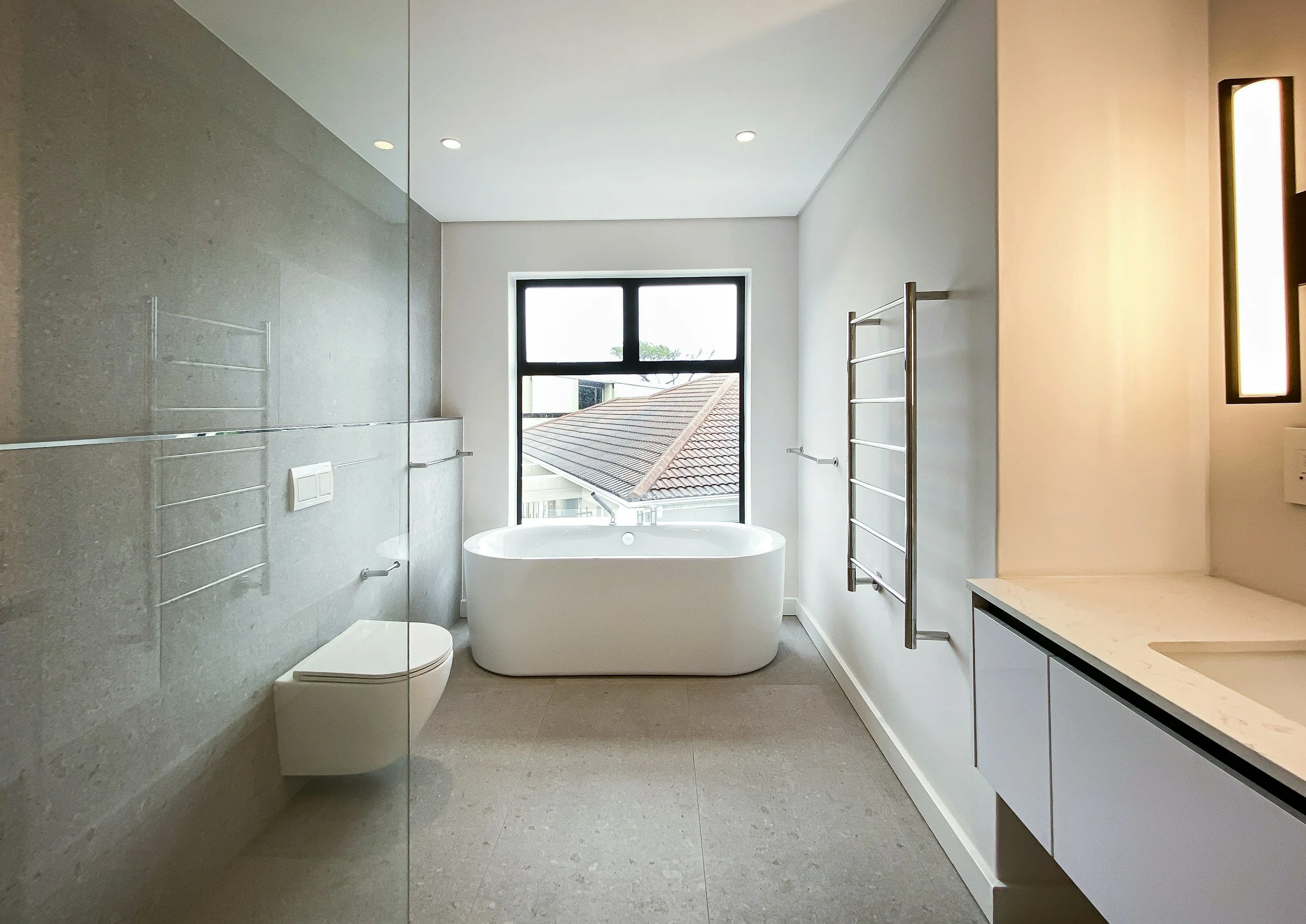 Modern bathroom with a white bathtub near a window, towel racks, a wall-mounted toilet, and a marble countertop sink.