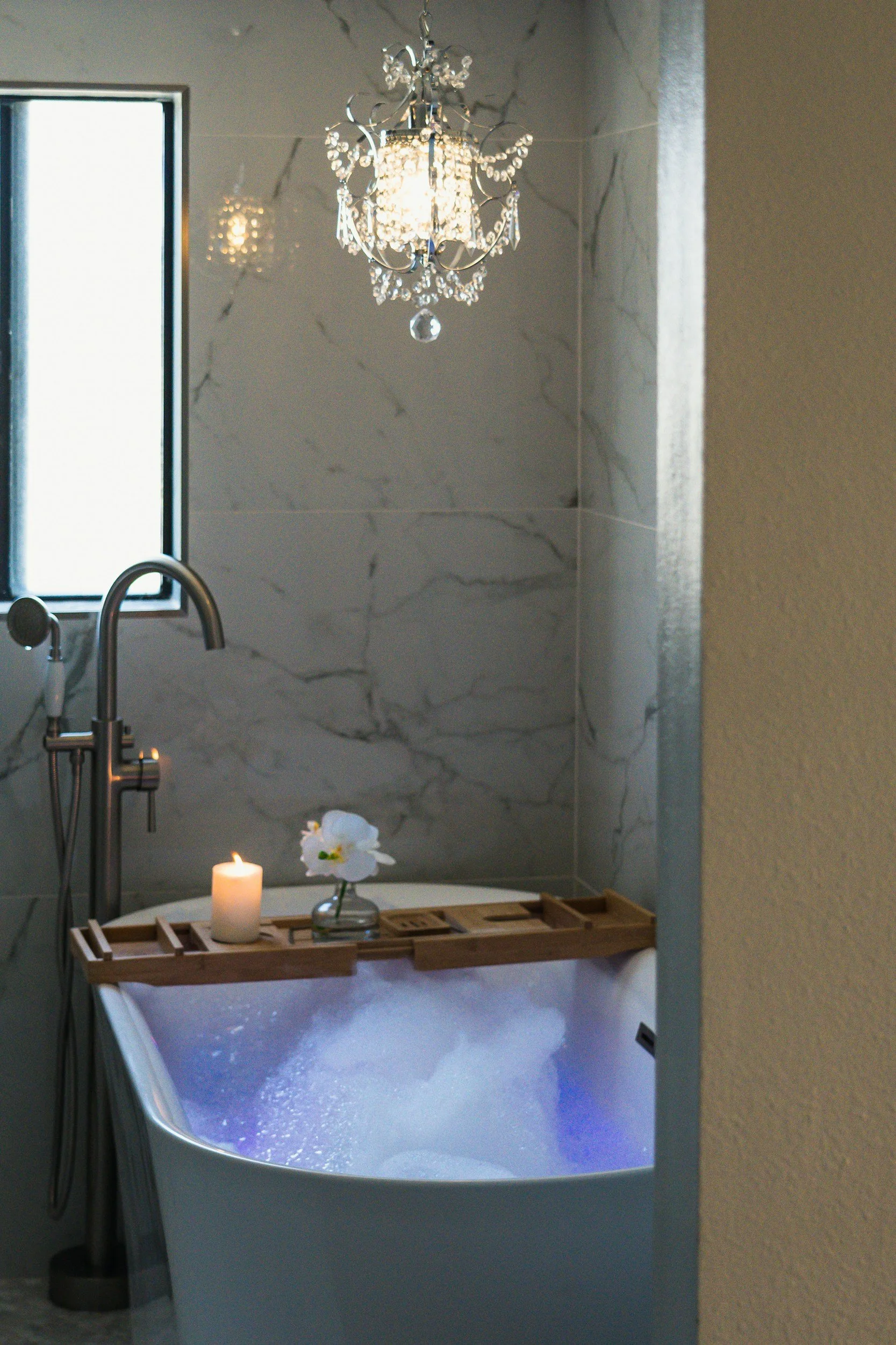 A bathroom with a bathtub filled with water, a floating candle, and an orchid flower in a vase on a wooden tray, a window, a chandelier, and marble walls.