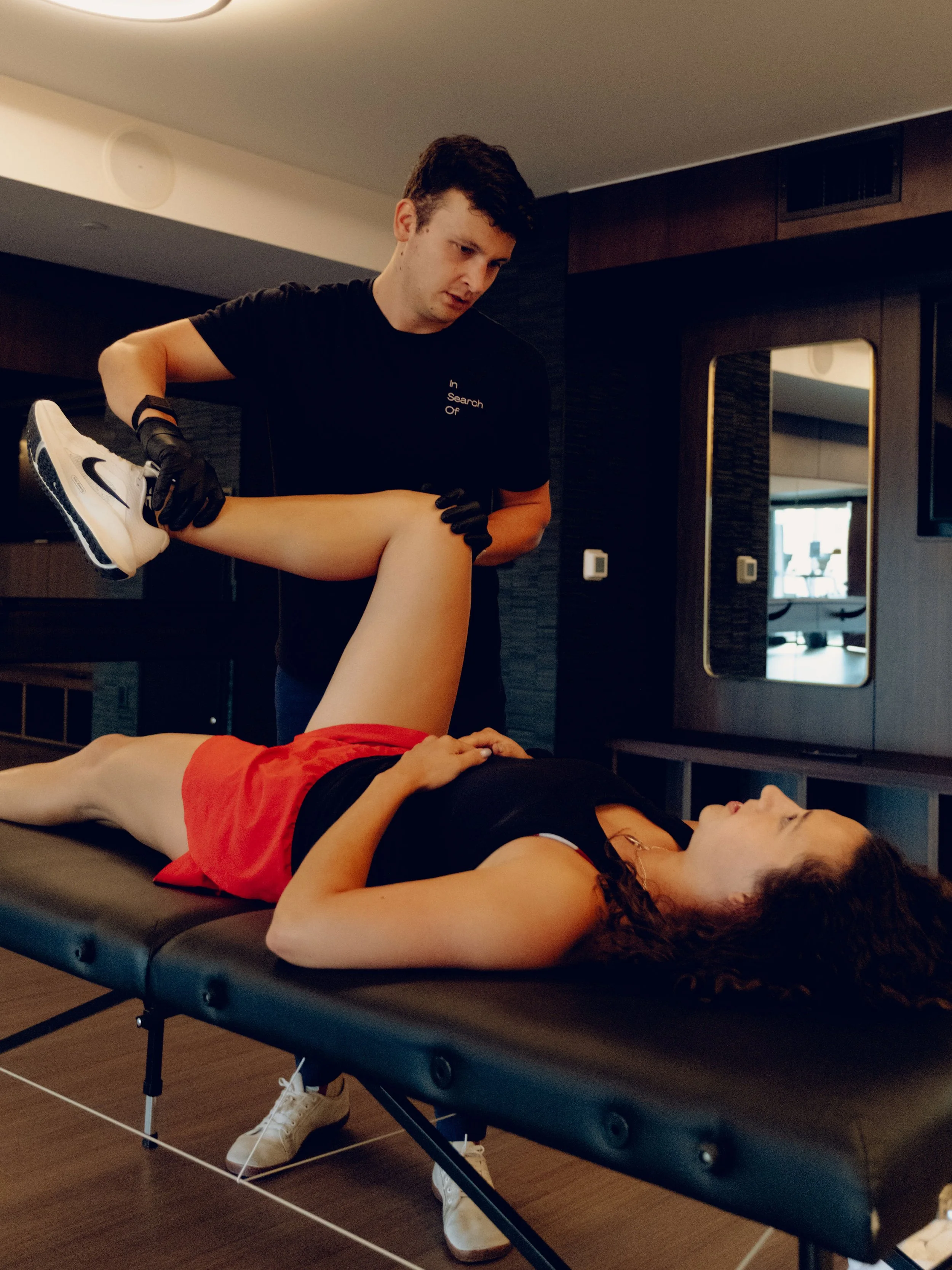 A physical therapist in black gloves assisting a woman with a leg stretch on a black therapy table in a therapy room.