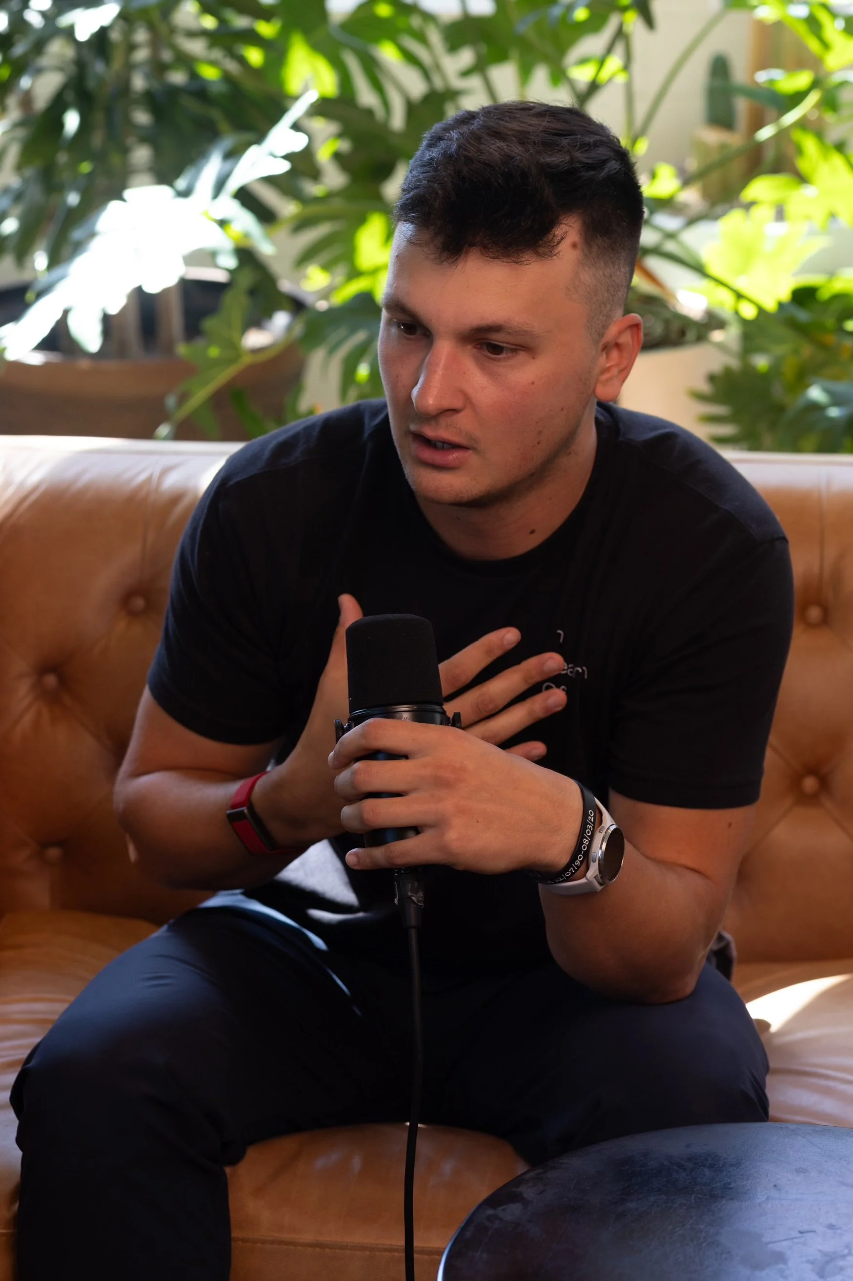 A young man sitting on a leather couch, speaking into a microphone, with lush green plants in the background.