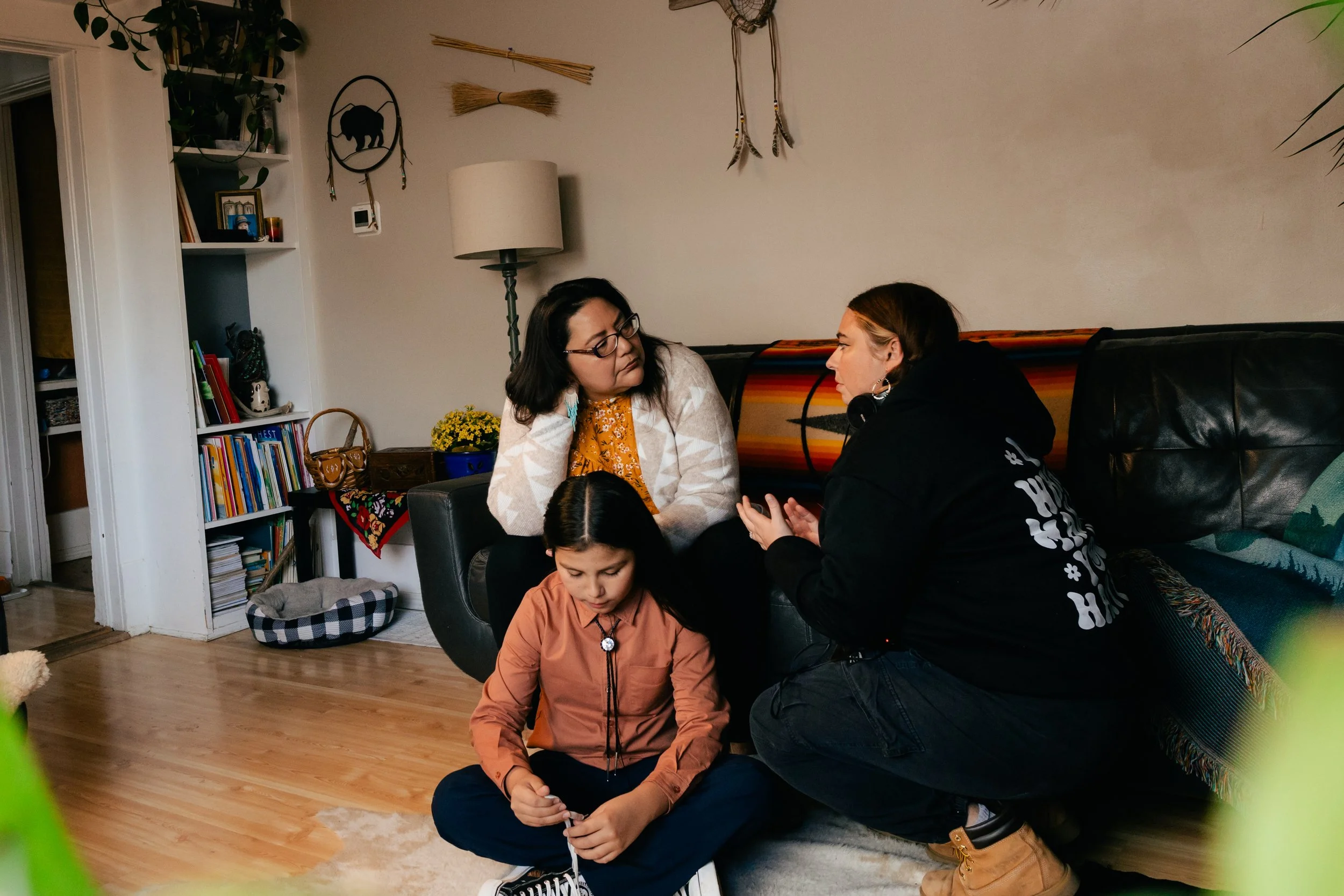 Three people, two women and one girl, sit and kneel on the floor in a cozy living room, engaging in conversation.