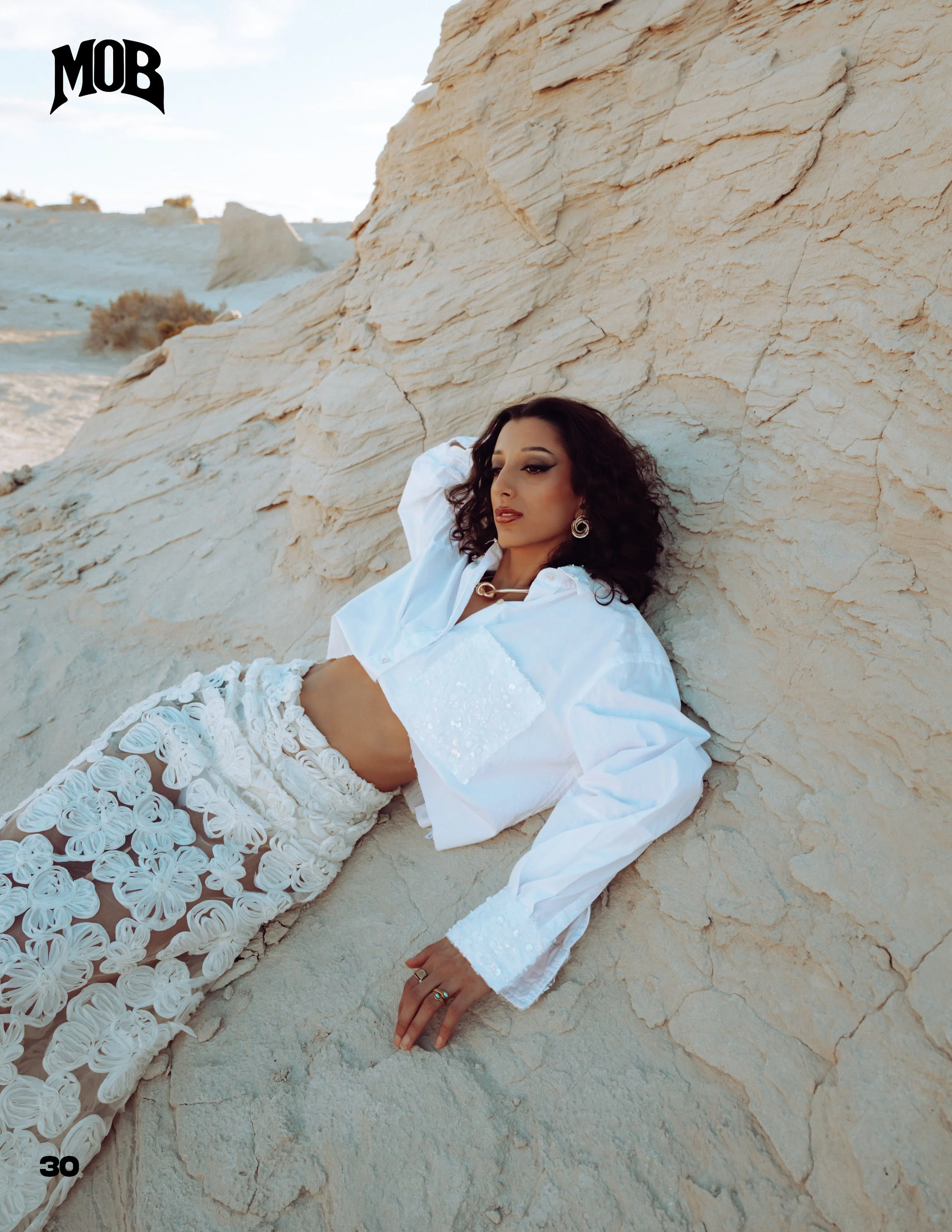A woman with curly dark hair, wearing a white shirt and a floral white skirt, lying against a large beige rock formation in a desert-like landscape.