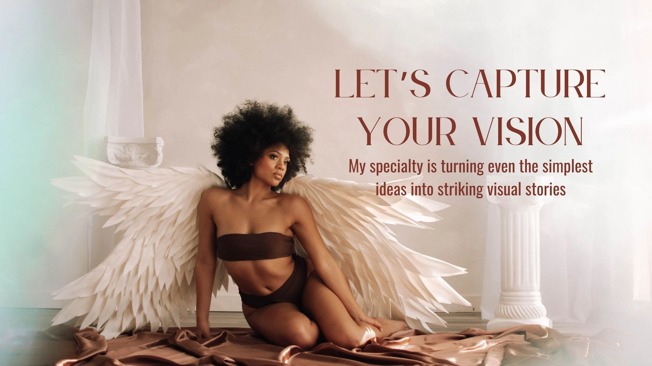 A woman with large white feathered angel wings kneels on a satin sheet in front of a white wall with classical columns. She is wearing a brown strapless top and matching bottoms and looks thoughtfully to the side. Taken by Emmie Edmo