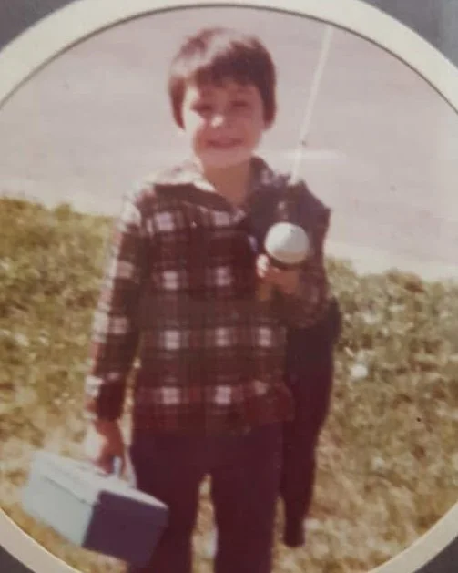 A young boy with short dark hair, smiling, holding a small white ball in one hand and a silver lunchbox in the other, outdoors on a grassy area with clear sky. Emmie Edmo's family