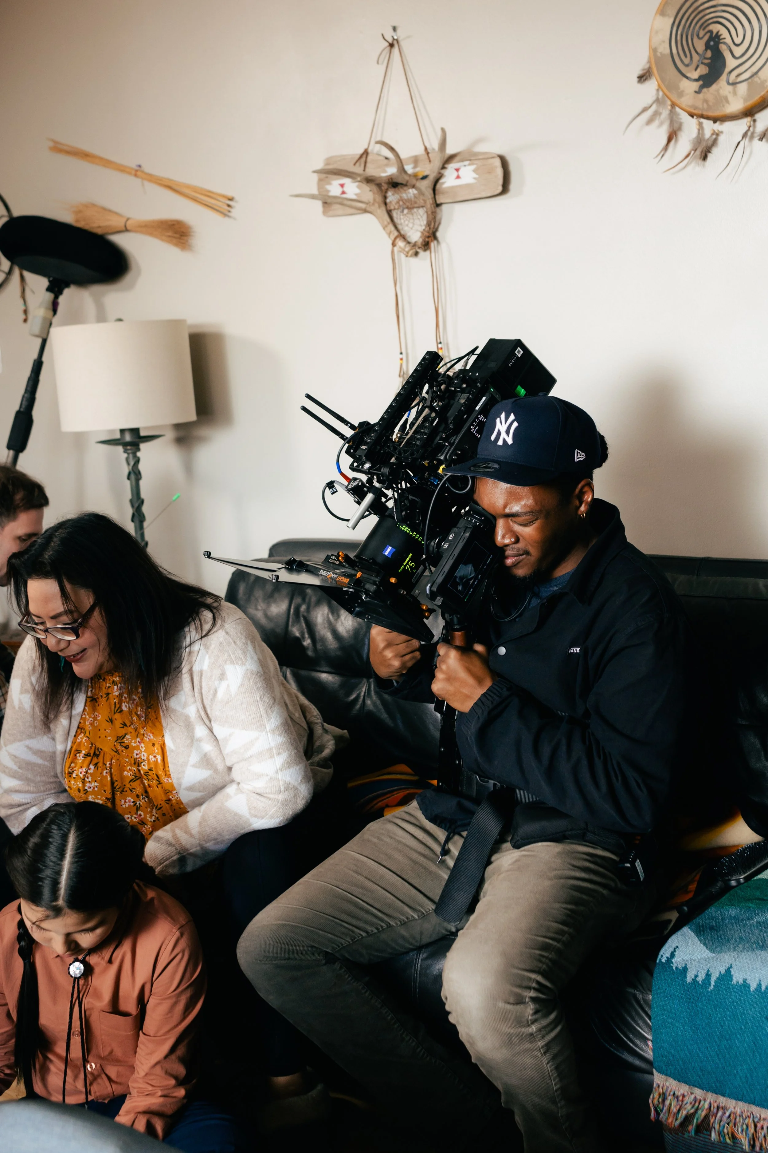 A man filming with a professional camera on a sofa in a living room with Native American decorative items on the wall.