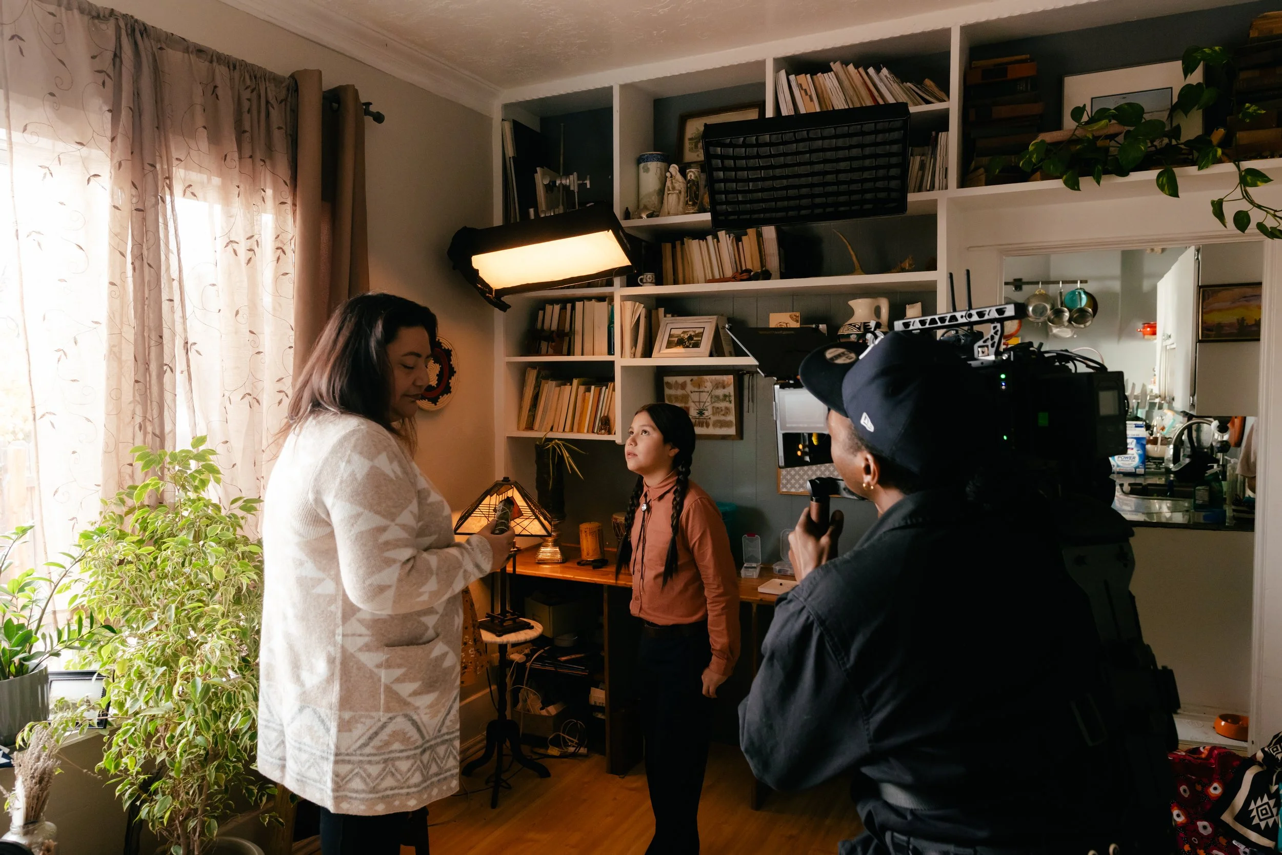 A woman in a white patterned coat appears to be holding a phone while talking in a living room, with another woman in a brown jacket listening. A person operating video equipment is recording the scene. The room has shelves with books, plants, and va