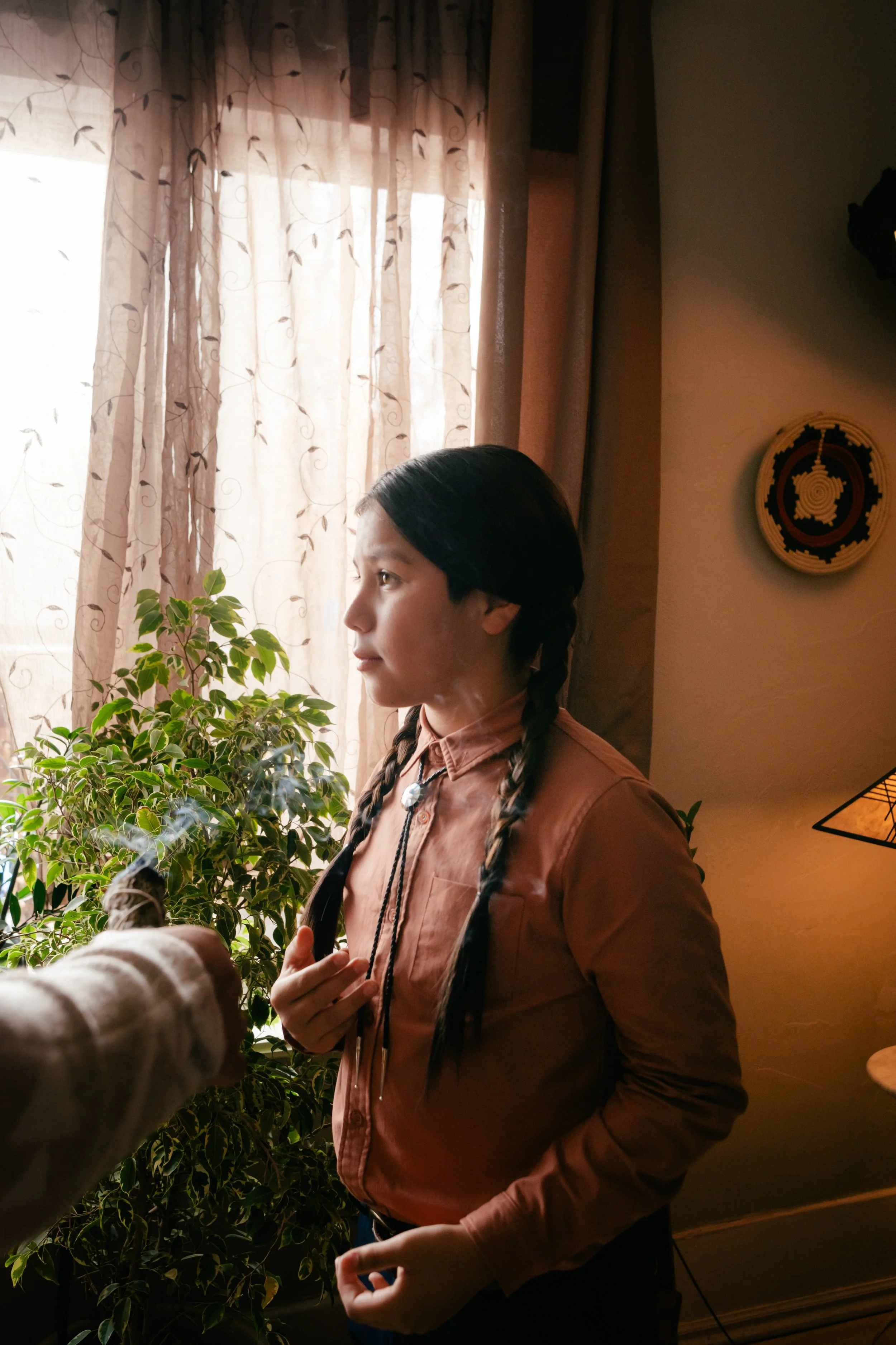 A woman with braided hair and a peach-colored shirt standing indoors near a window with beige curtains, surrounded by plants.