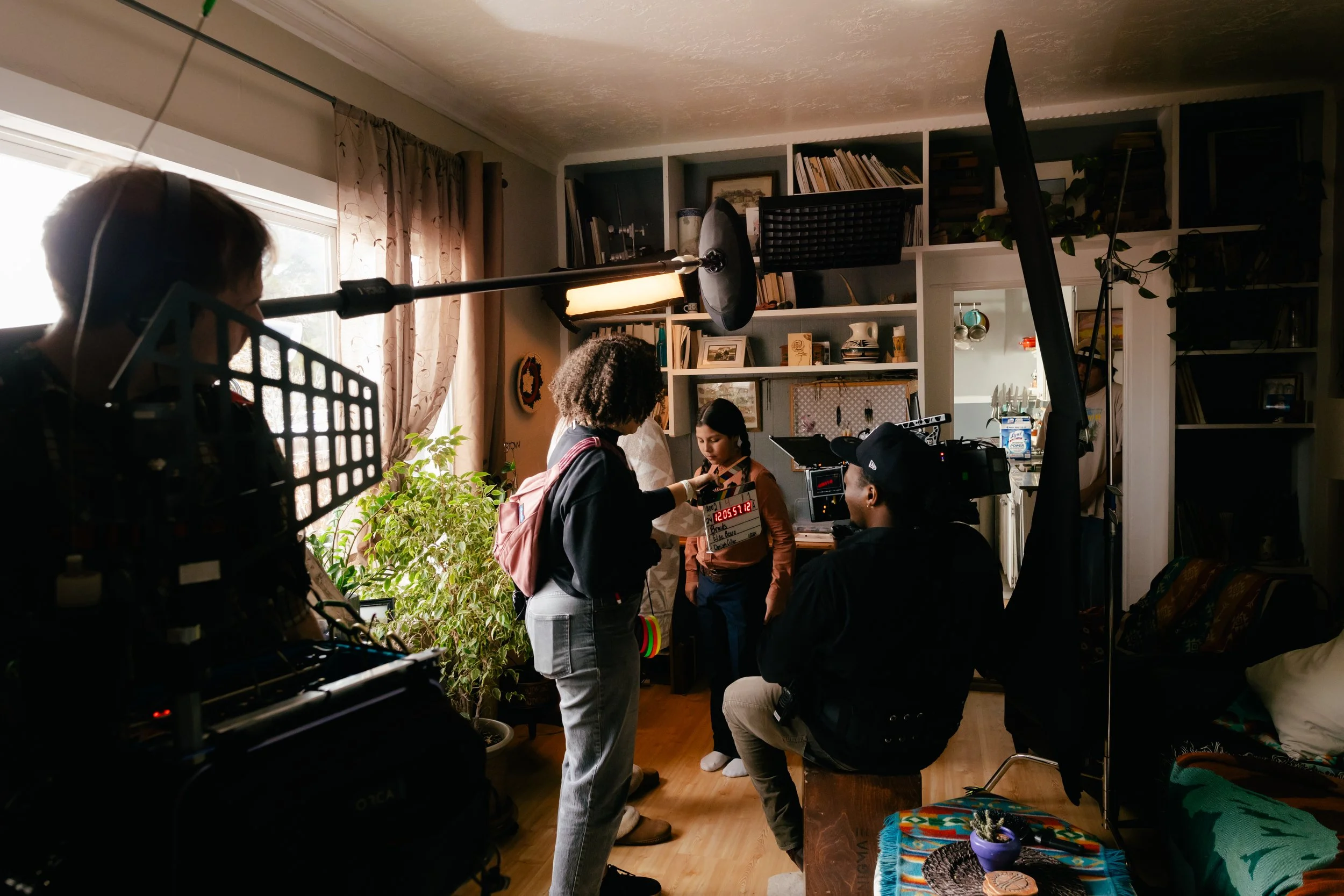 Film crew filming inside a cozy living room with shelves, plants, and natural light, capturing an interview or dialogue with a young woman.