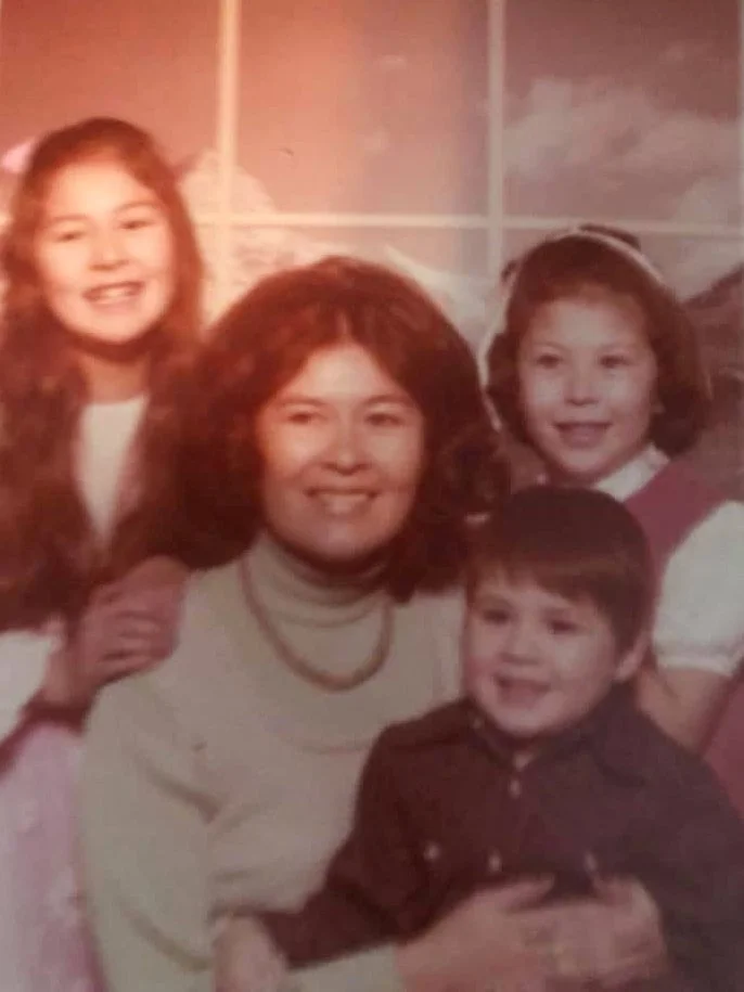A woman with curly hair and three children, all smiling, in front of a painted backdrop of a window and mountains. Emmie Edmo's family