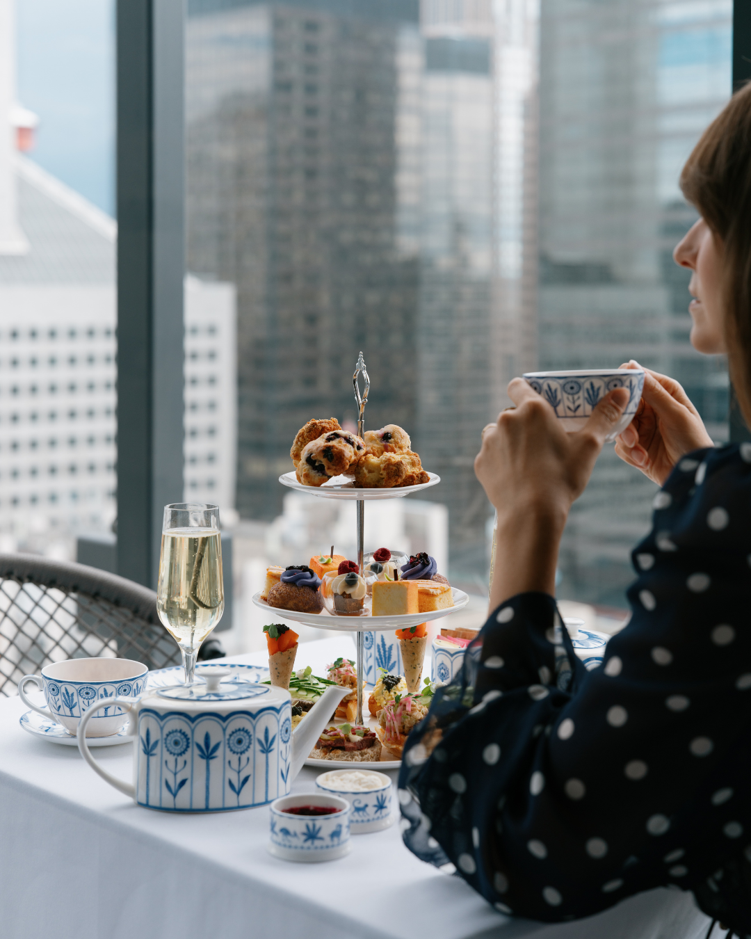 A woman enjoying tea with a tiered stand of assorted desserts, including scones, cakes, and pastries, set on a table with a city skyline view in the background.