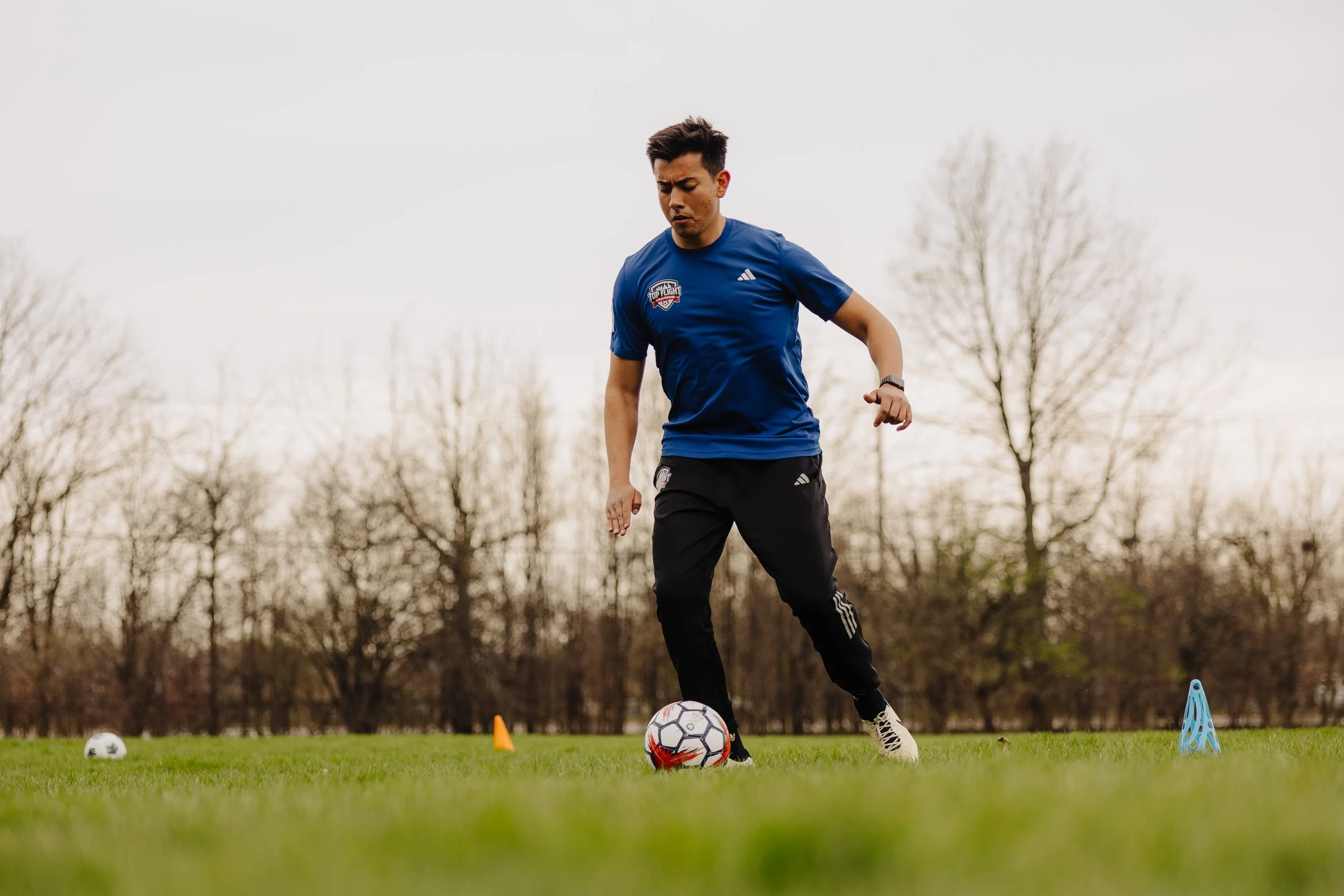 Man practicing soccer outdoors on a grassy field with training cones and a ball.