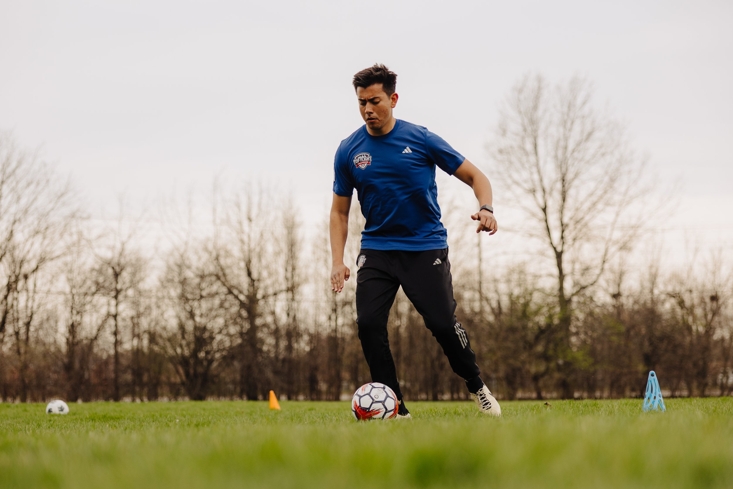 A person standing on a grassy field, dressed in soccer gear, preparing to kick a soccer ball with their right foot. They are wearing black shorts with a 'Top Flight Training' logo, cream Nike soccer cleats, and black and white striped socks. The background shows a few trees and a cloudy sky.