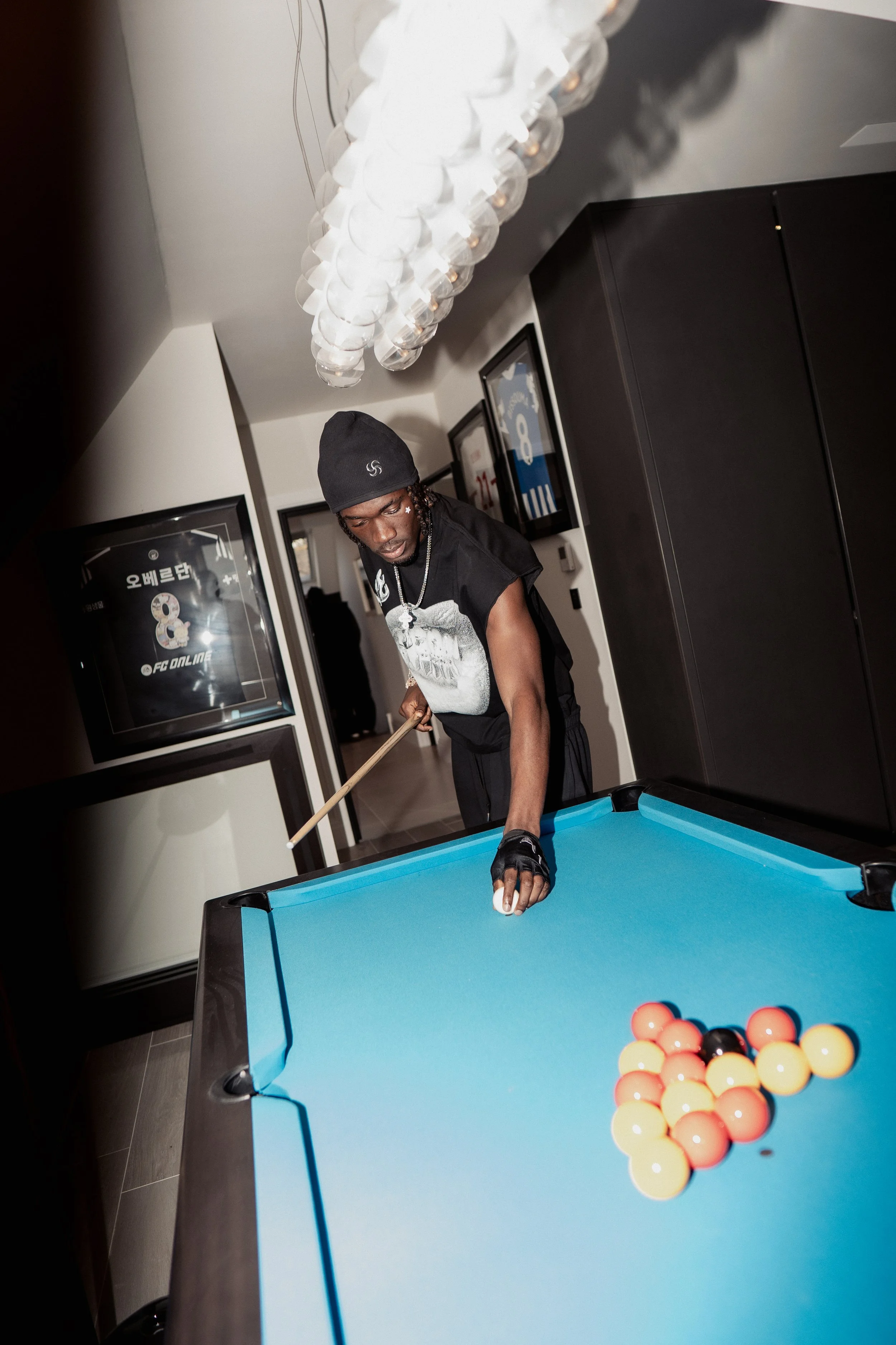 Un homme joue au billard dans une pièce moderne, portant un bonnet noir, un T-shirt noir avec un dessin, et une main gantée, avec un éclairage moderne et des affiches encadrées au mur.