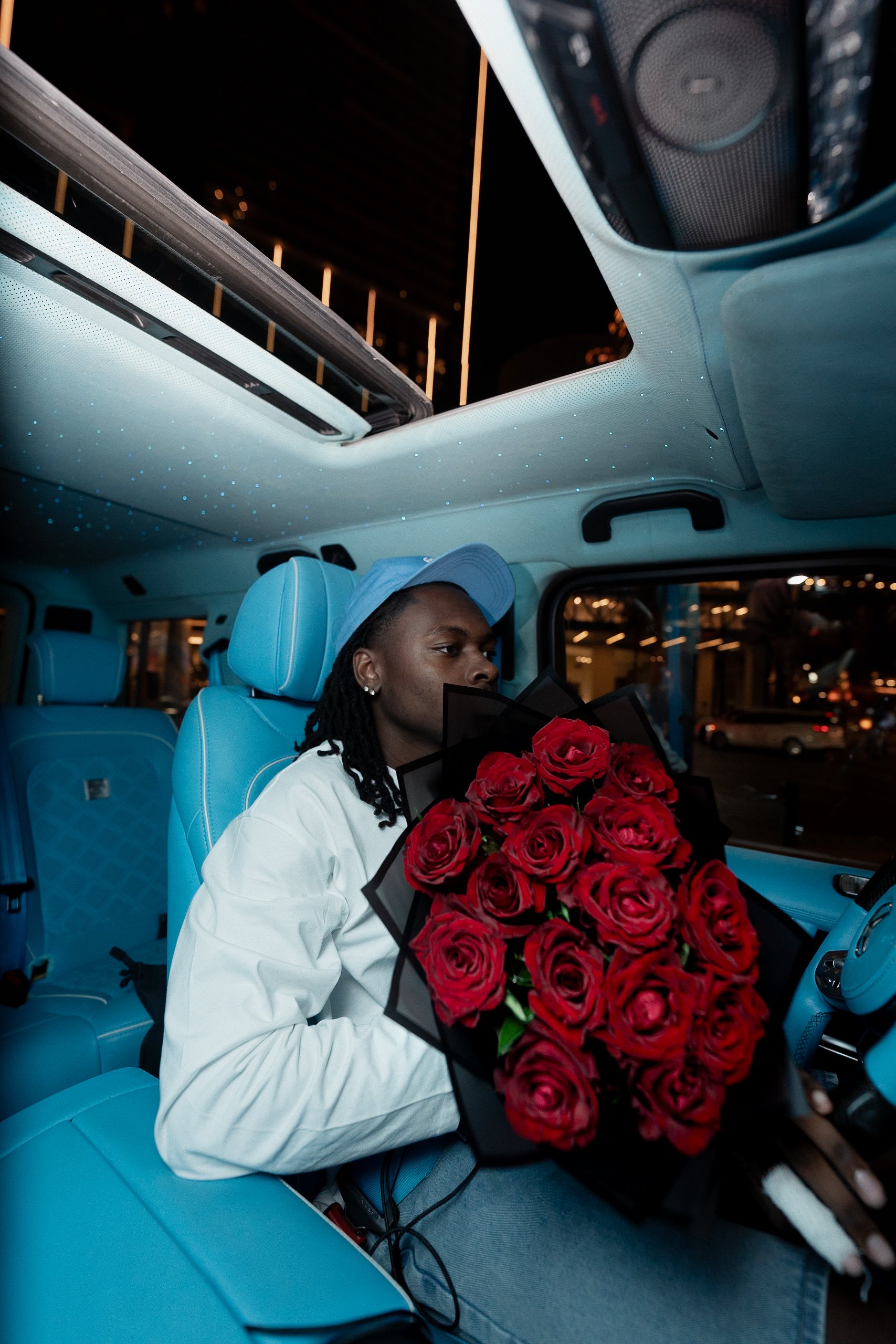 Une personne assise dans un véhicule, tenant un bouquet de roses rouges, portant une casquette bleue et une veste blanche, à l'intérieur d'une voiture lumineuse et moderne.