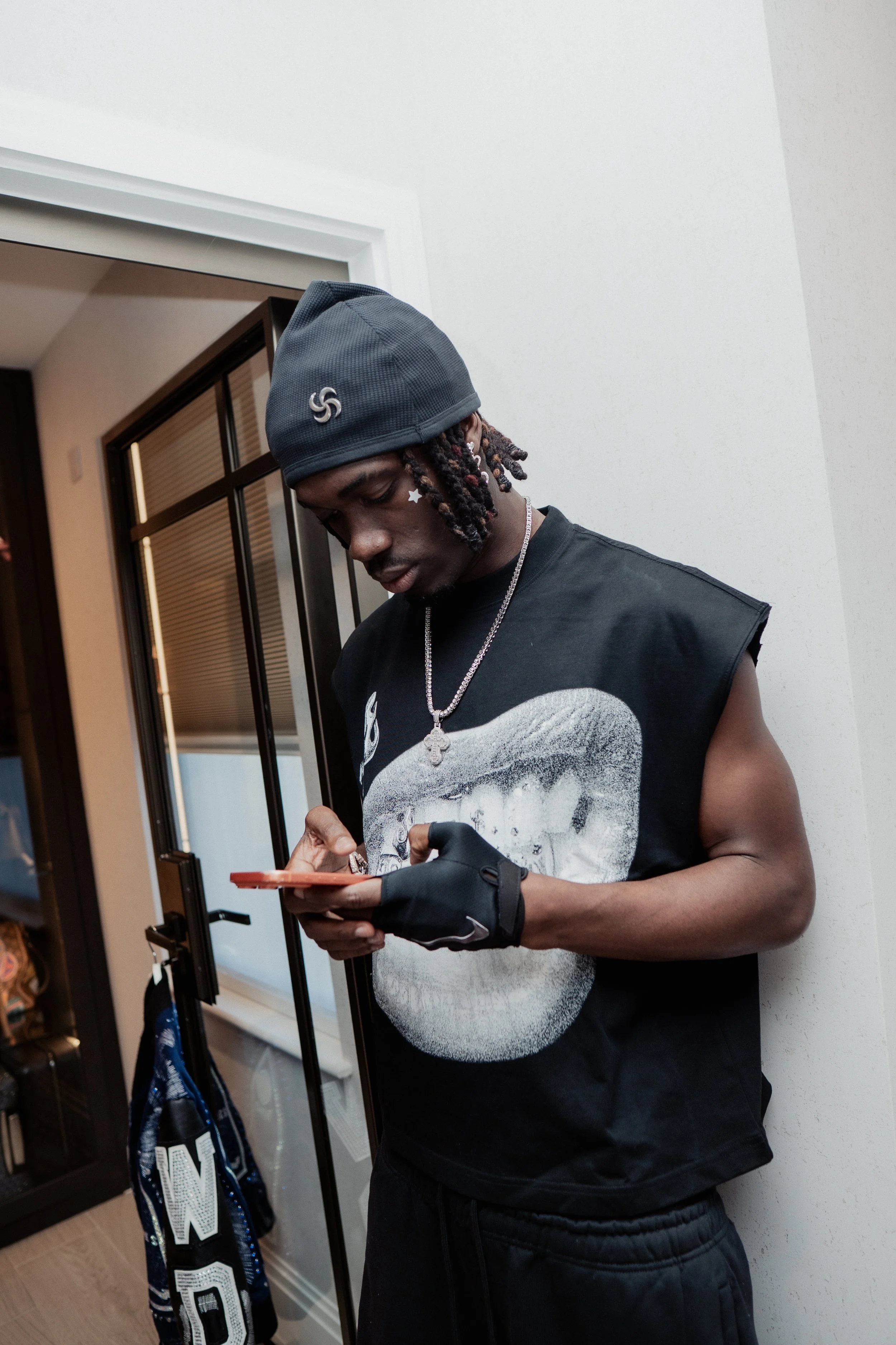 Un jeune homme noir portant un bonnet gris, un bracelet noir, un t-shirt sans manches avec une image de visage, une chaîne en argent avec un pendentif, semble regarder son téléphone dans une pièce intérieure.