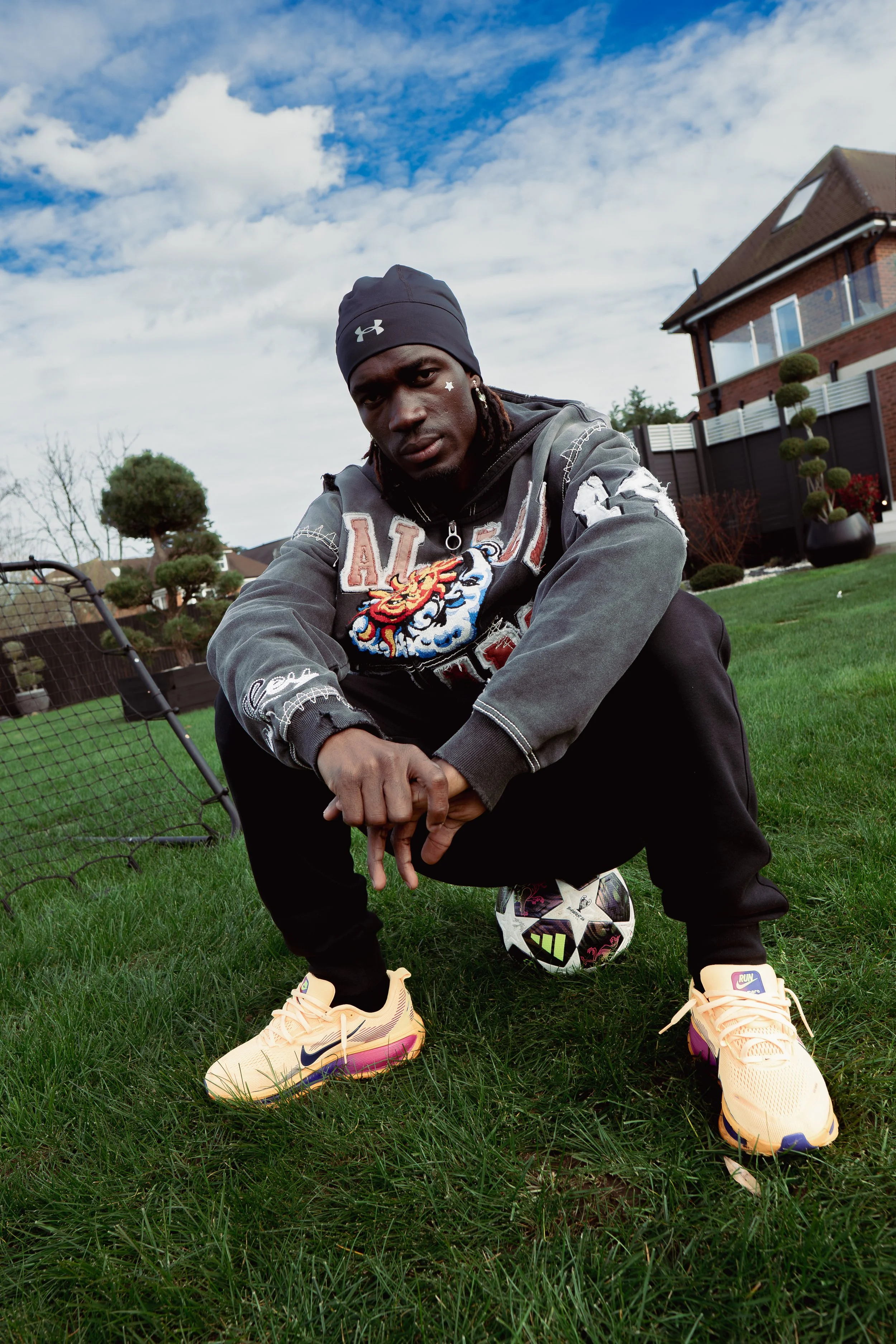 Un jeune homme en tenue décontractée, portant un bonnet noir et des baskets jaunes, est assis sur le gazon avec un ballon de football à ses pieds. Il regarde l'objectif avec une expression sérieuse dans un jardin résidentiel avec des arbres et une ma