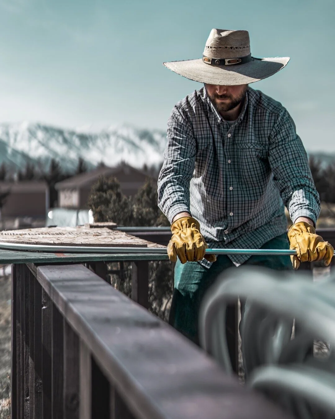 A person using a homemade hoop bender to shape low tunnel hoops from electrical conduit, wearing a wide-brimmed hat with snowy mountains blurred in the background.