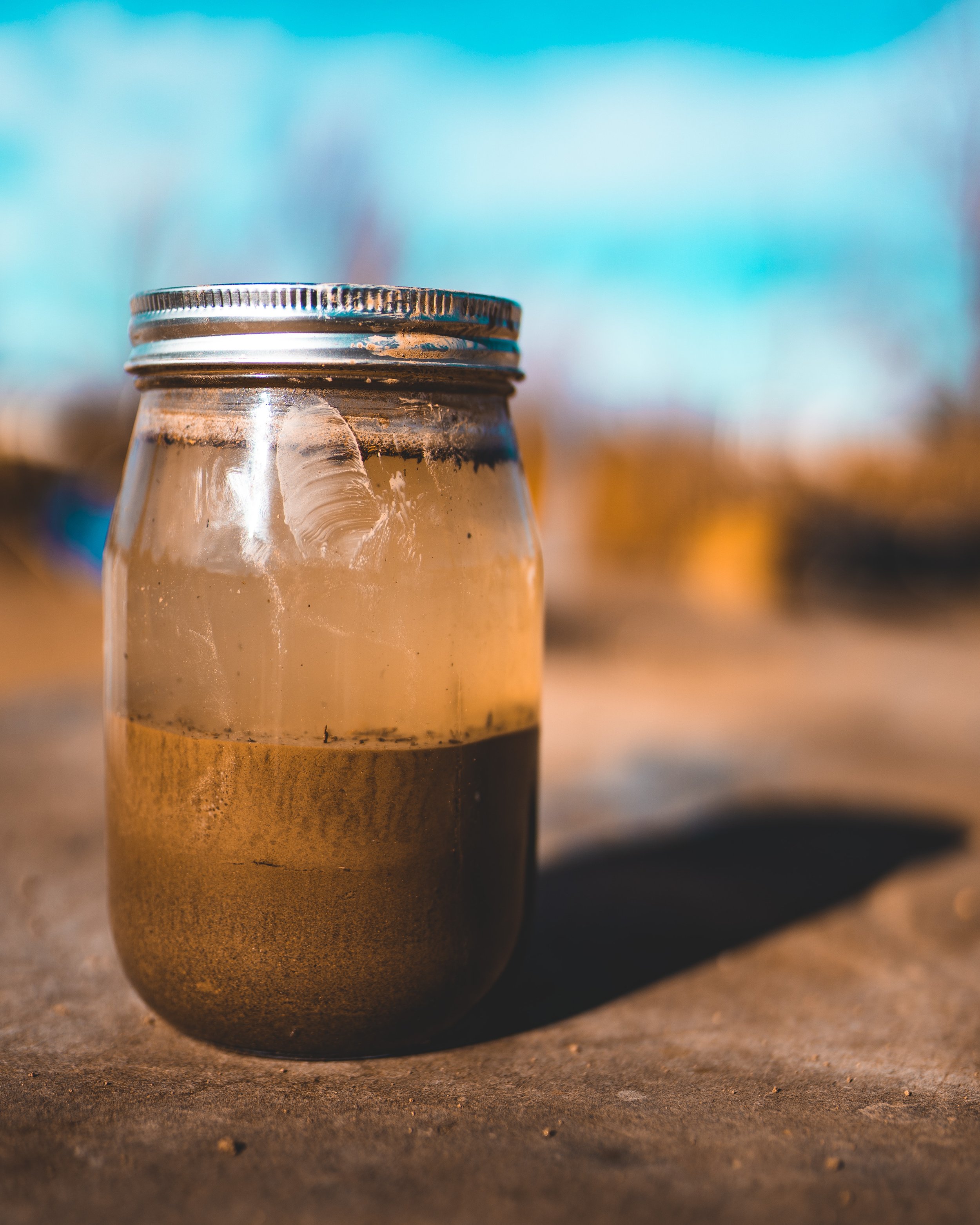 Mason jar filled with soil and water for a jar test to assess soil composition, placed on an outdoor bench with a blue sky in the background.