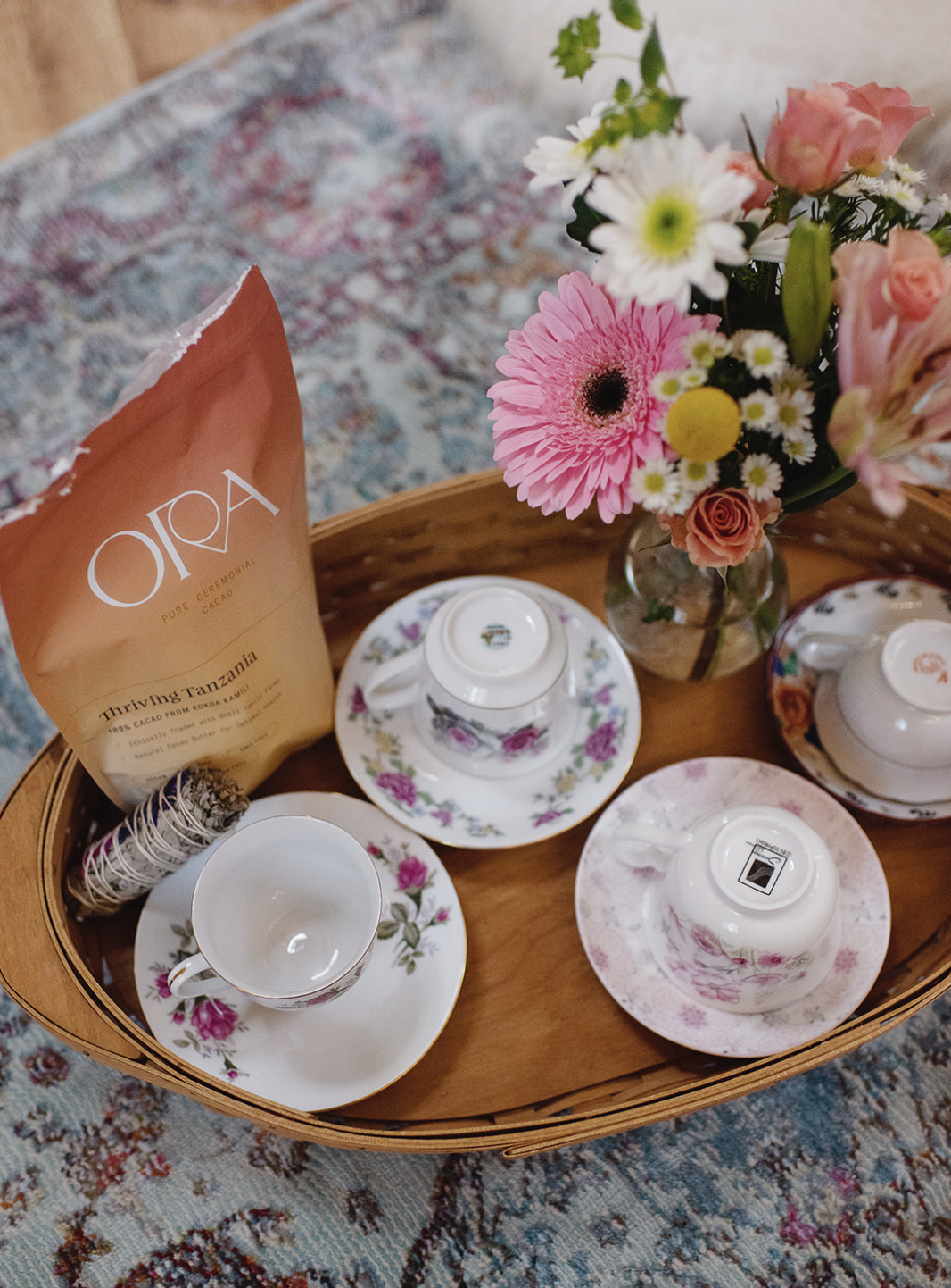 Tea set with floral cups and saucers, a carton of Oira cacao, a bouquet of colorful flowers in a glass vase on a wooden tray, placed on a patterned rug.