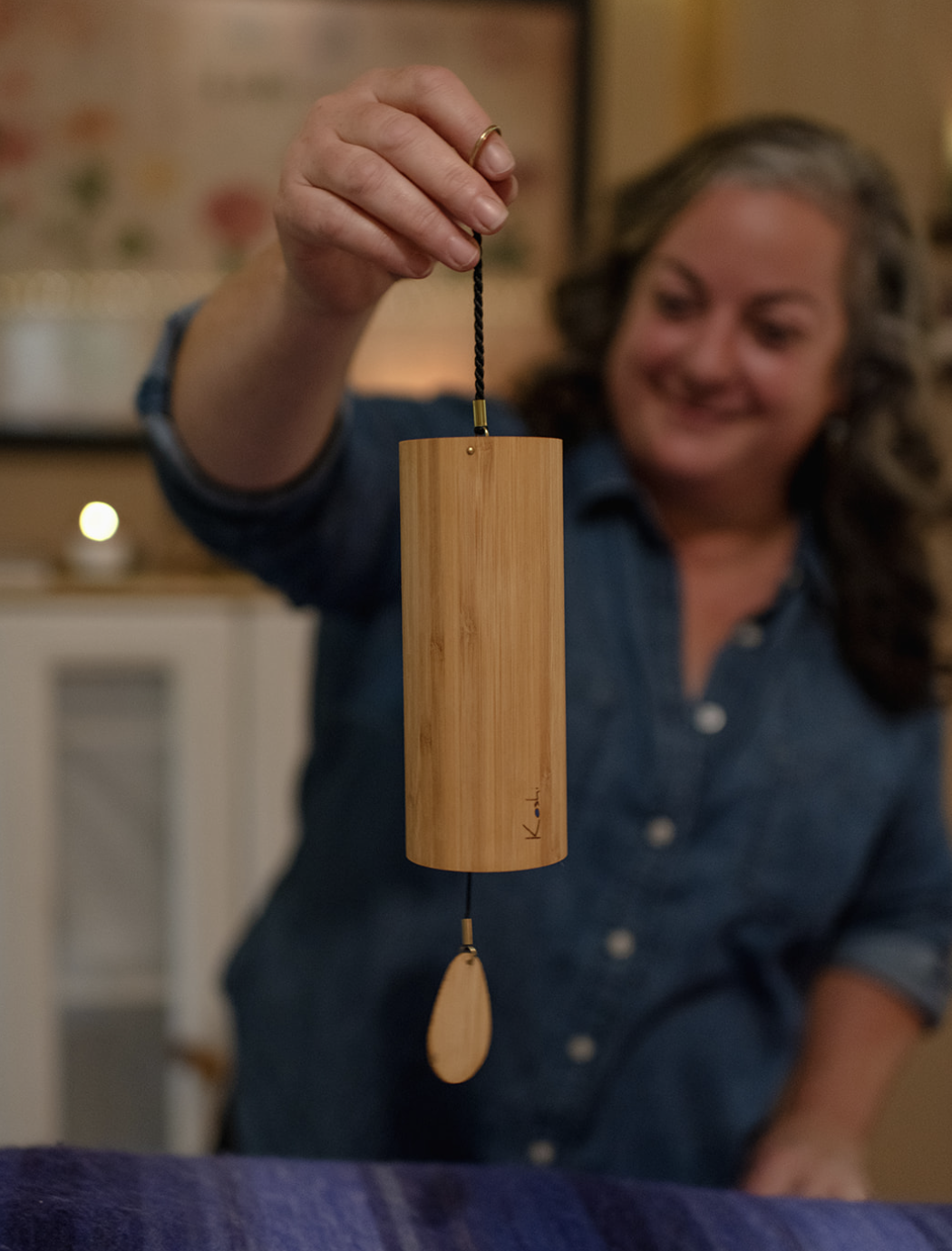 Woman holding a wooden wind chime with a black cord and a wooden teardrop-shaped weight, smiling in a cozy indoor setting.