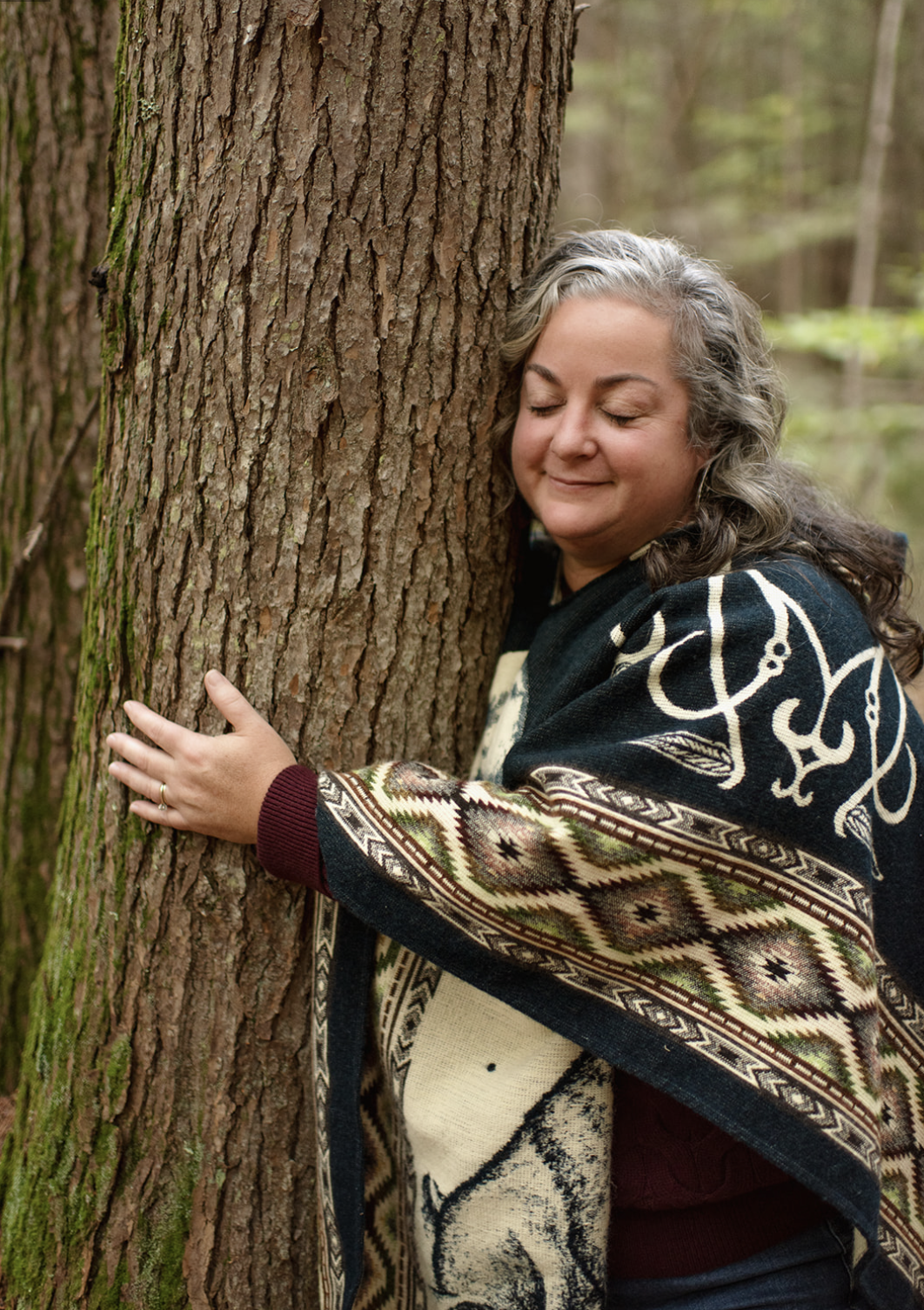 A woman hugging a tree in a forest with her eyes closed and a peaceful expression.
