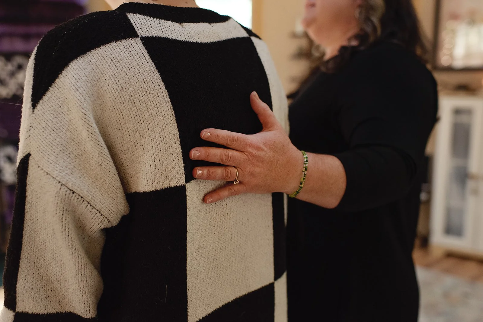 A woman in a black top and green bracelet places her hand on the shoulder of a person wearing a black and white checkered sweater.