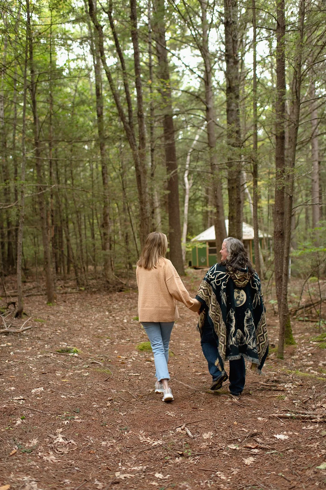 Two women walking hand in hand on a dirt path through a forest with tall trees and a small green and white building in the background.