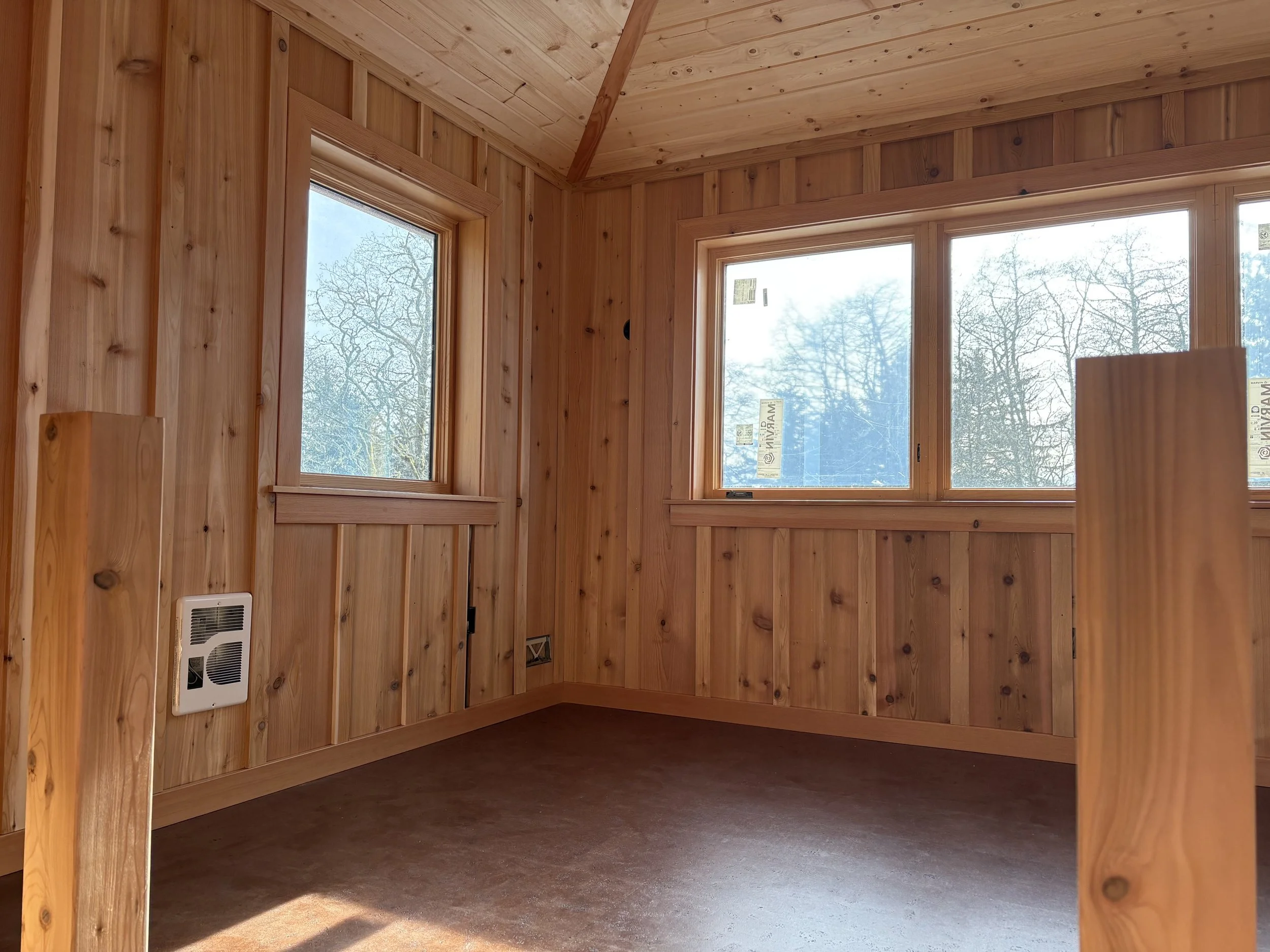 Interior of a wooden room with natural wood panel walls and ceiling, two large windows showing leafless trees and a clear sky, and a vent on the lower wall.