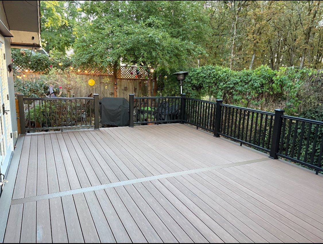 Empty outdoor deck with black railing, surrounded by trees and greenery, with potted plants and string lights on the left side.