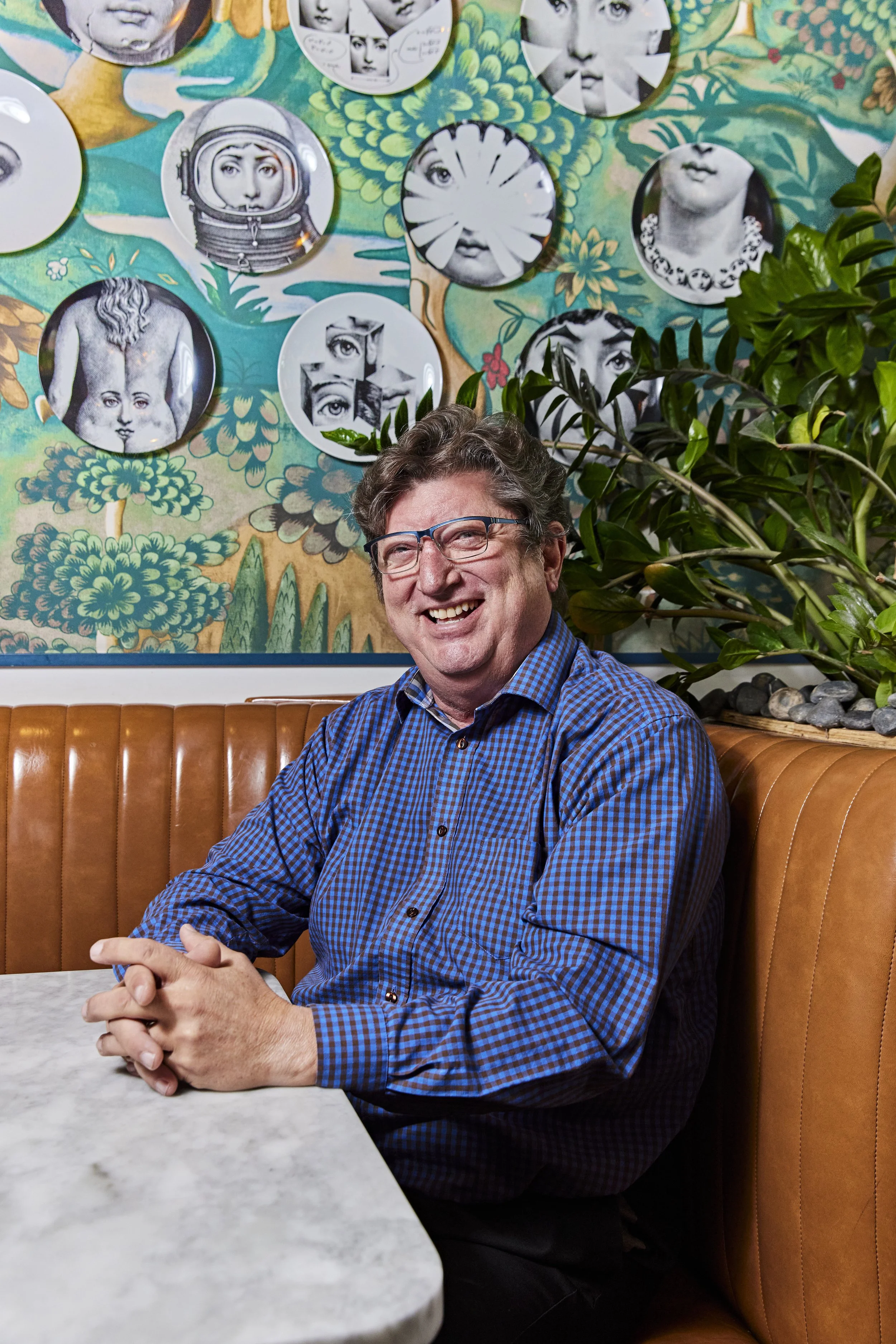 A smiling man with glasses, wearing a blue checkered shirt, sitting in a booth at a restaurant with a marble table. Behind him is a colorful wall with artwork featuring various black and white image collages and a large green leafy plant.