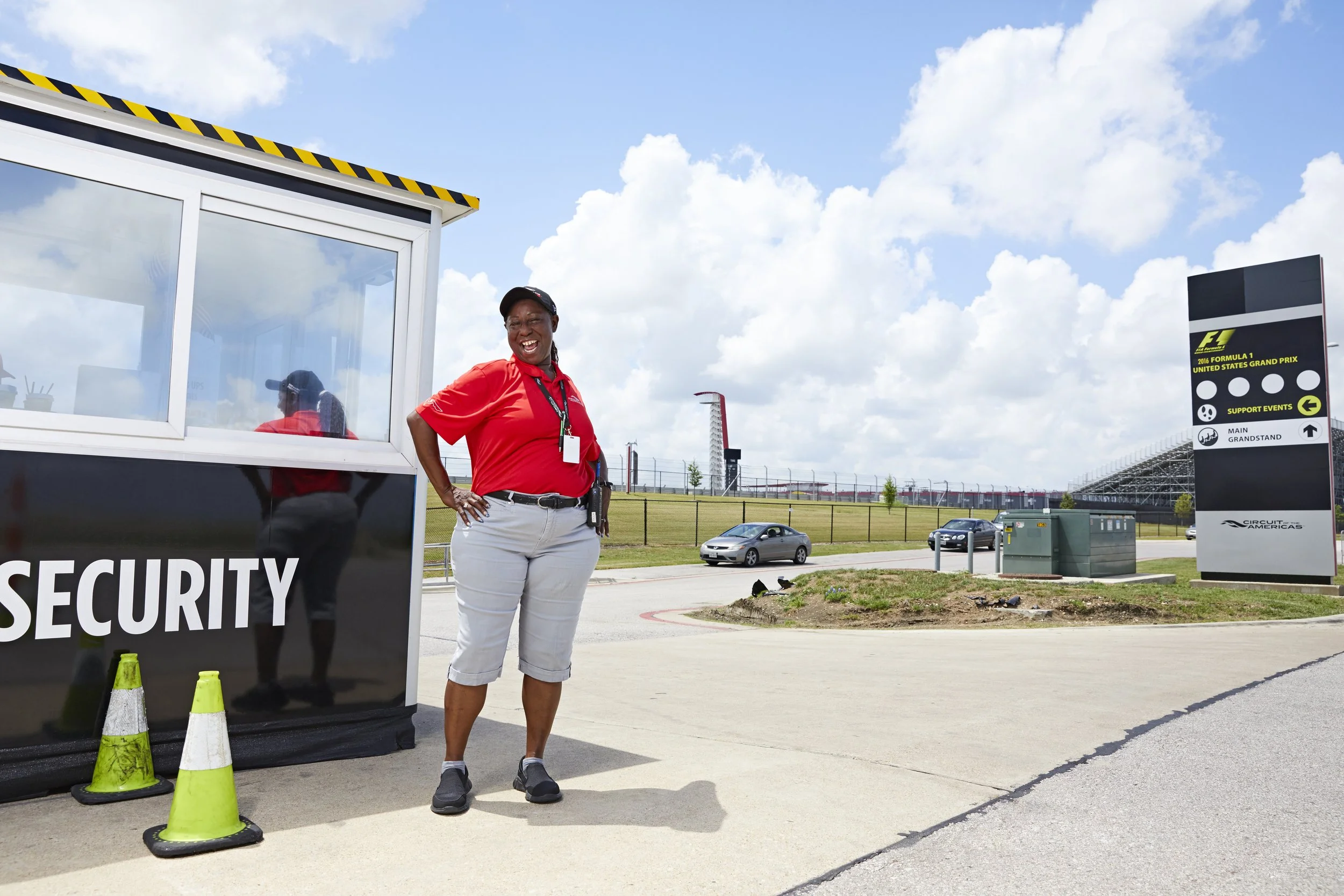 A woman in a red shirt and white pants standing near a security checkpoint at a racing event, with a Circuit of the Americas sign and cars in the background.