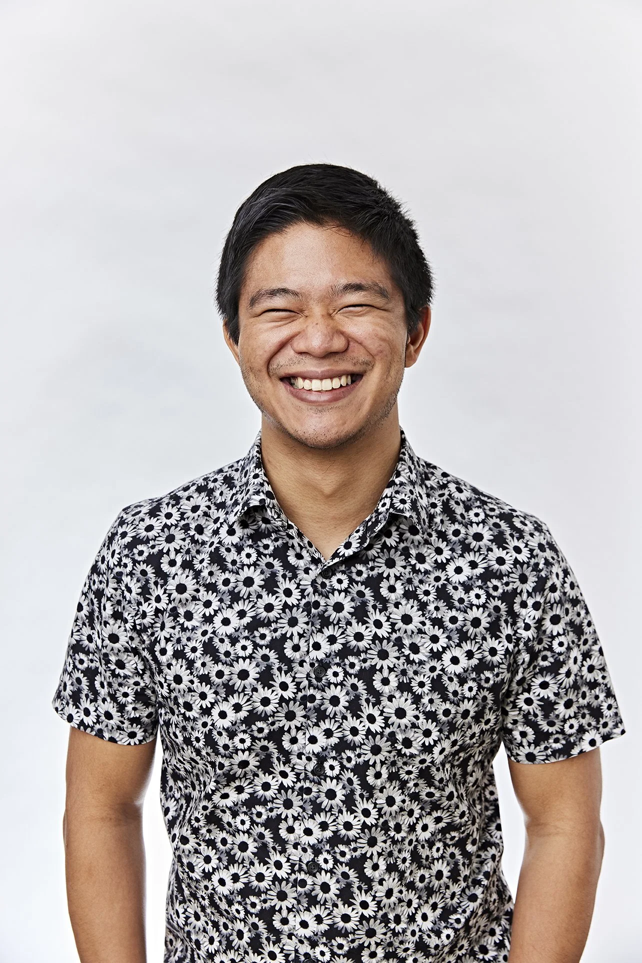 A smiling man with short black hair and facial hair, wearing a black and white floral shirt, standing against a plain white background.