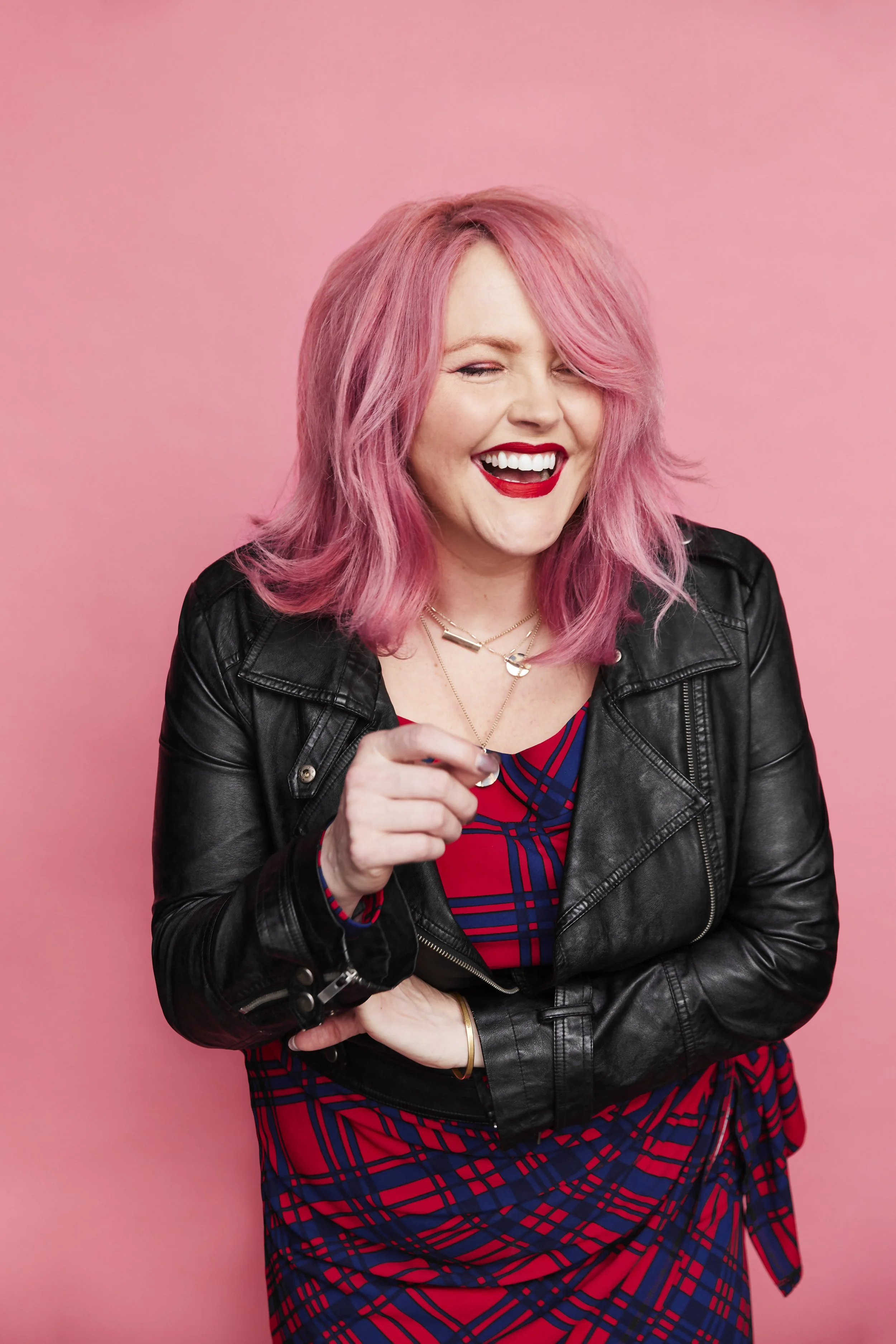 Woman with pink hair, smiling, wearing a black leather jacket and a red and blue plaid dress, standing against a pink background.