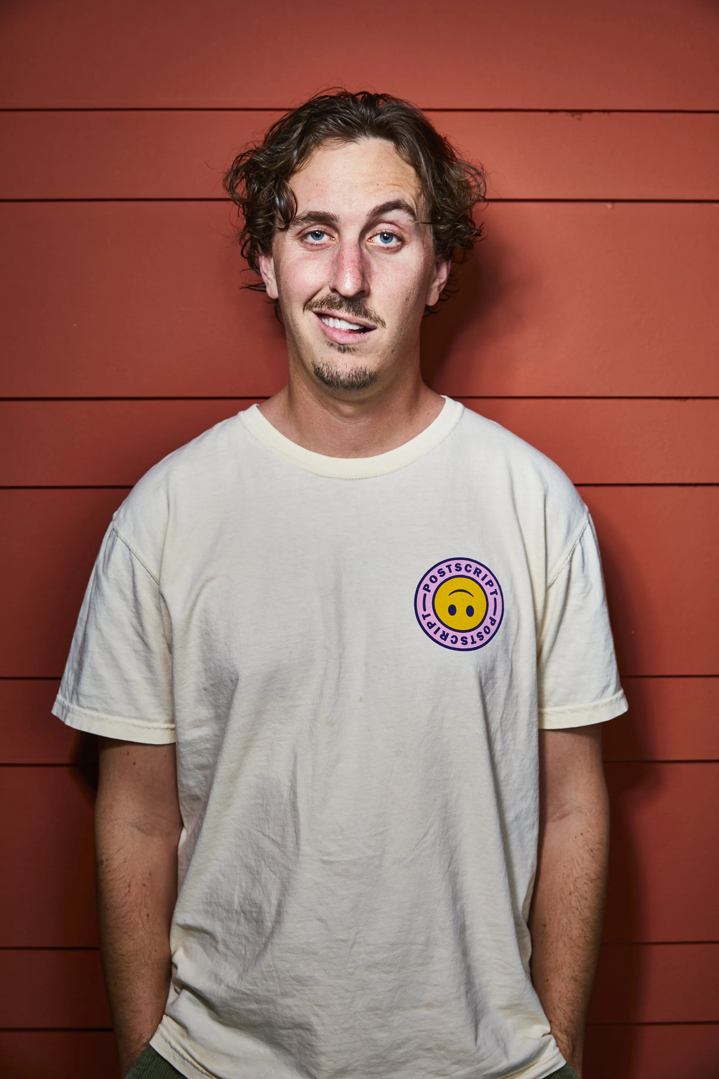 A young man with curly brown hair, blue eyes, and a light beard, standing against a red wooden wall. He is wearing a cream T-shirt with a round yellow smiley face logo and the word 'POSTSCRIPT' around it. He has a slight smirk and looks at the camera