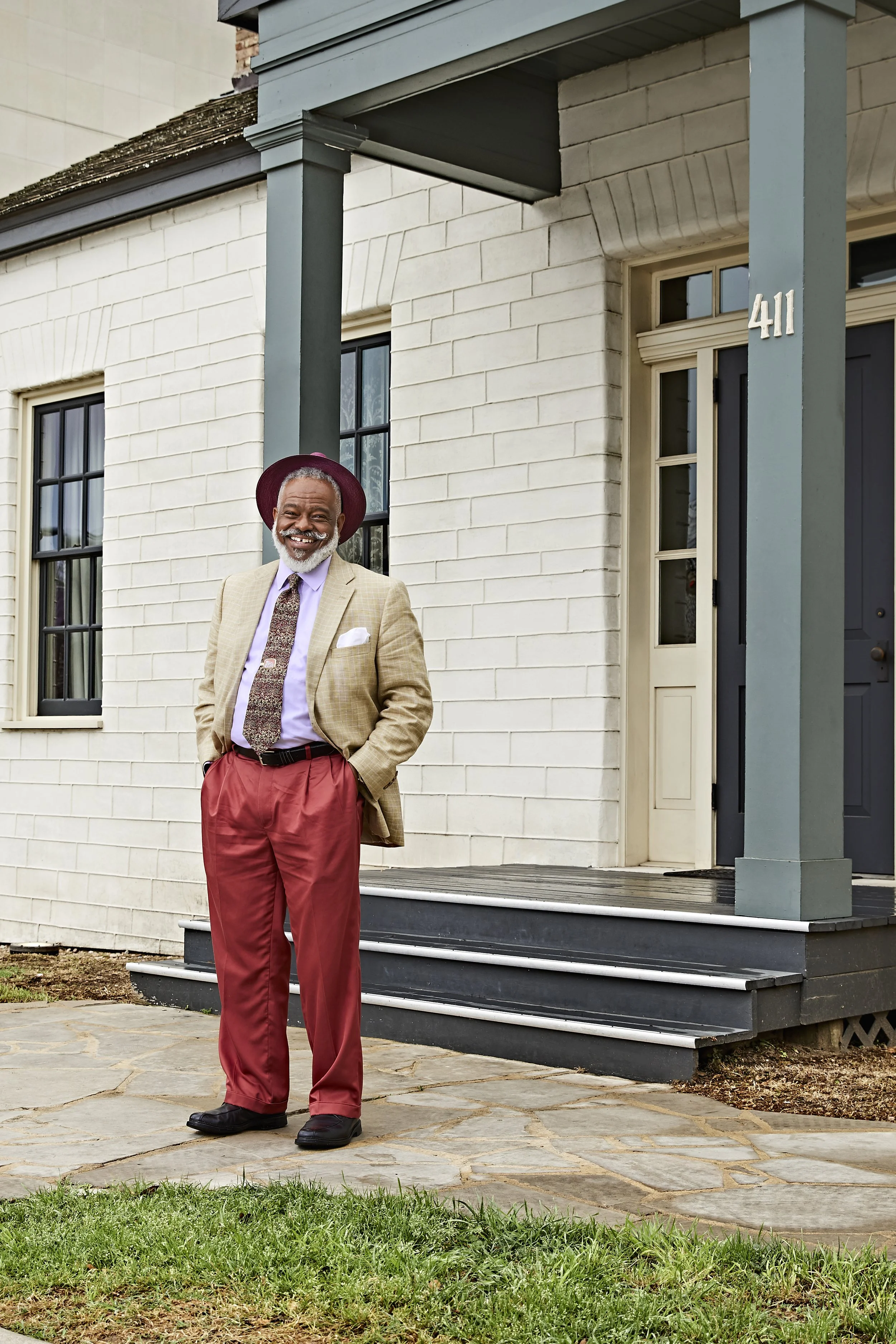 A smiling elderly man with a gray beard standing outside a white house, wearing a tan blazer, purple shirt, patterned tie, red pants, black shoes, and a burgundy hat.
