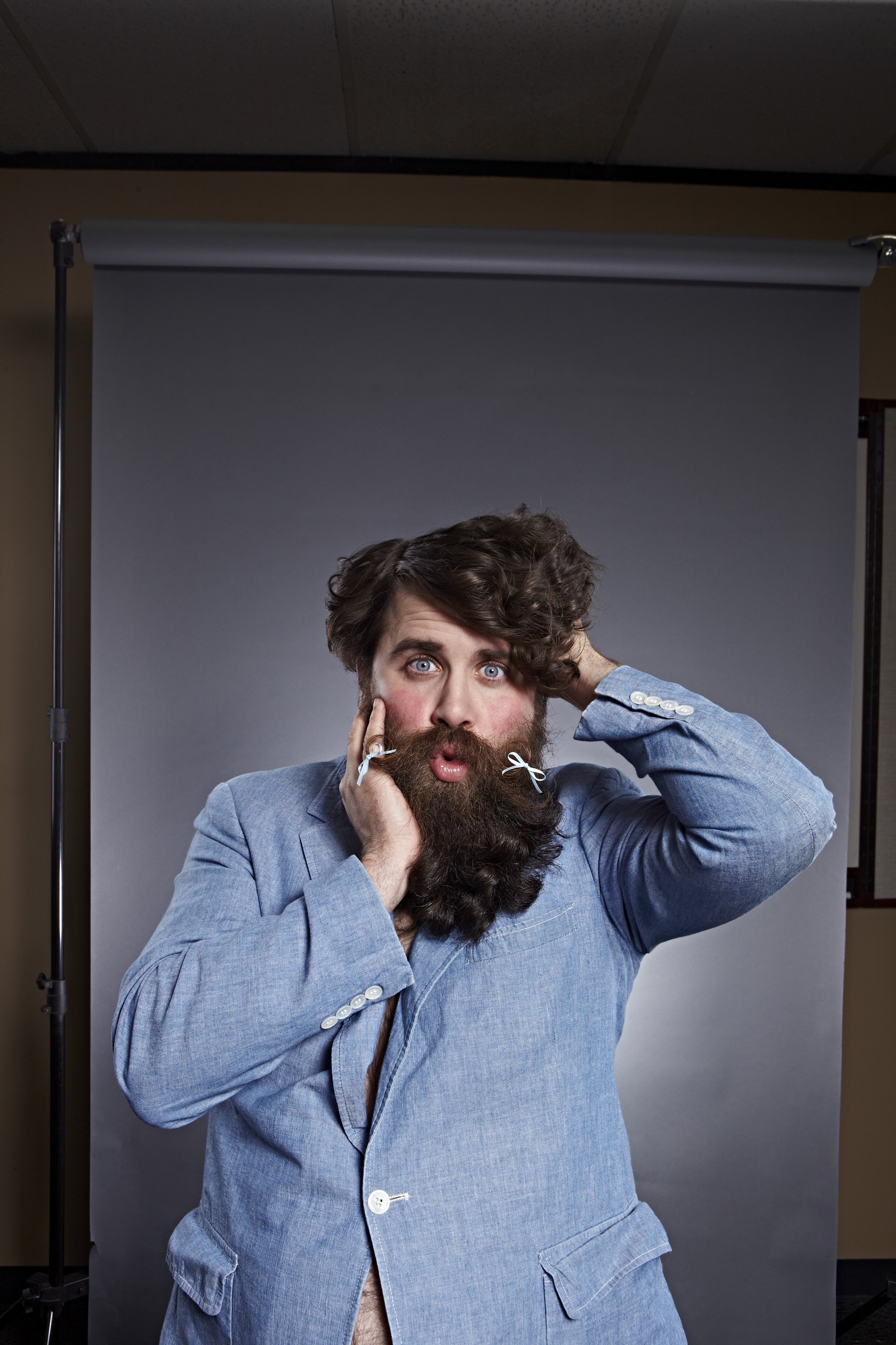 A man with a surprised expression, wearing a blue jacket, holding his face with one hand and touching his head with the other, standing in front of a gray backdrop.