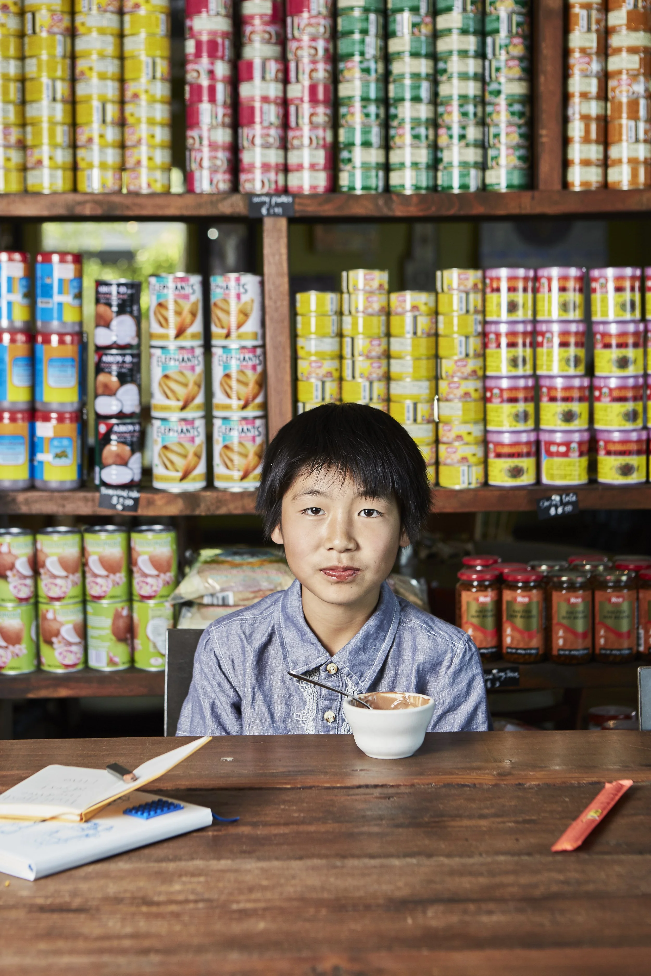 A young boy sitting at a wooden table with a bowl of chocolate ice cream in front of him, in a grocery store with shelves of canned goods behind him.
