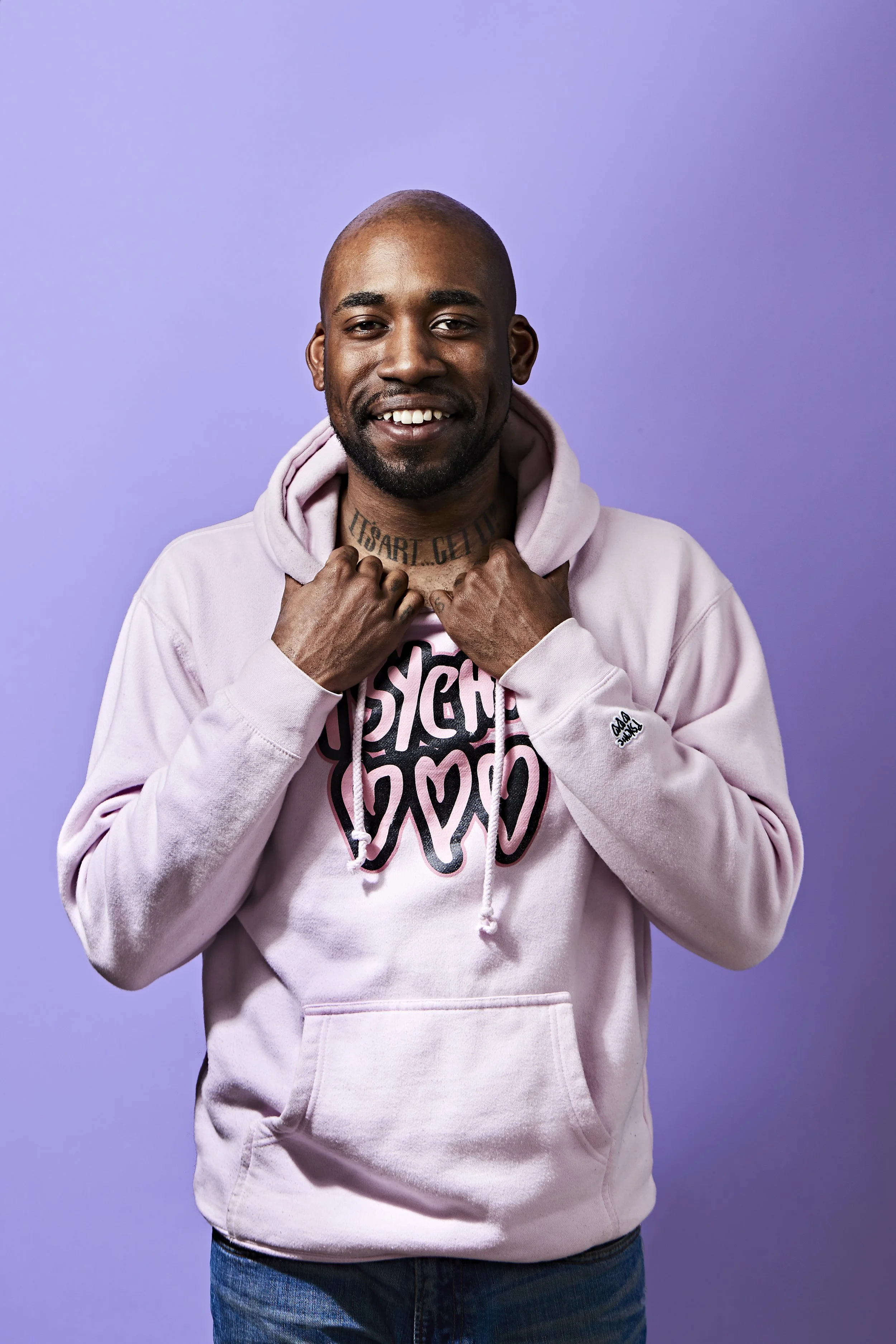 A smiling man wearing a light pink hoodie with text on the chest, standing against a purple background.