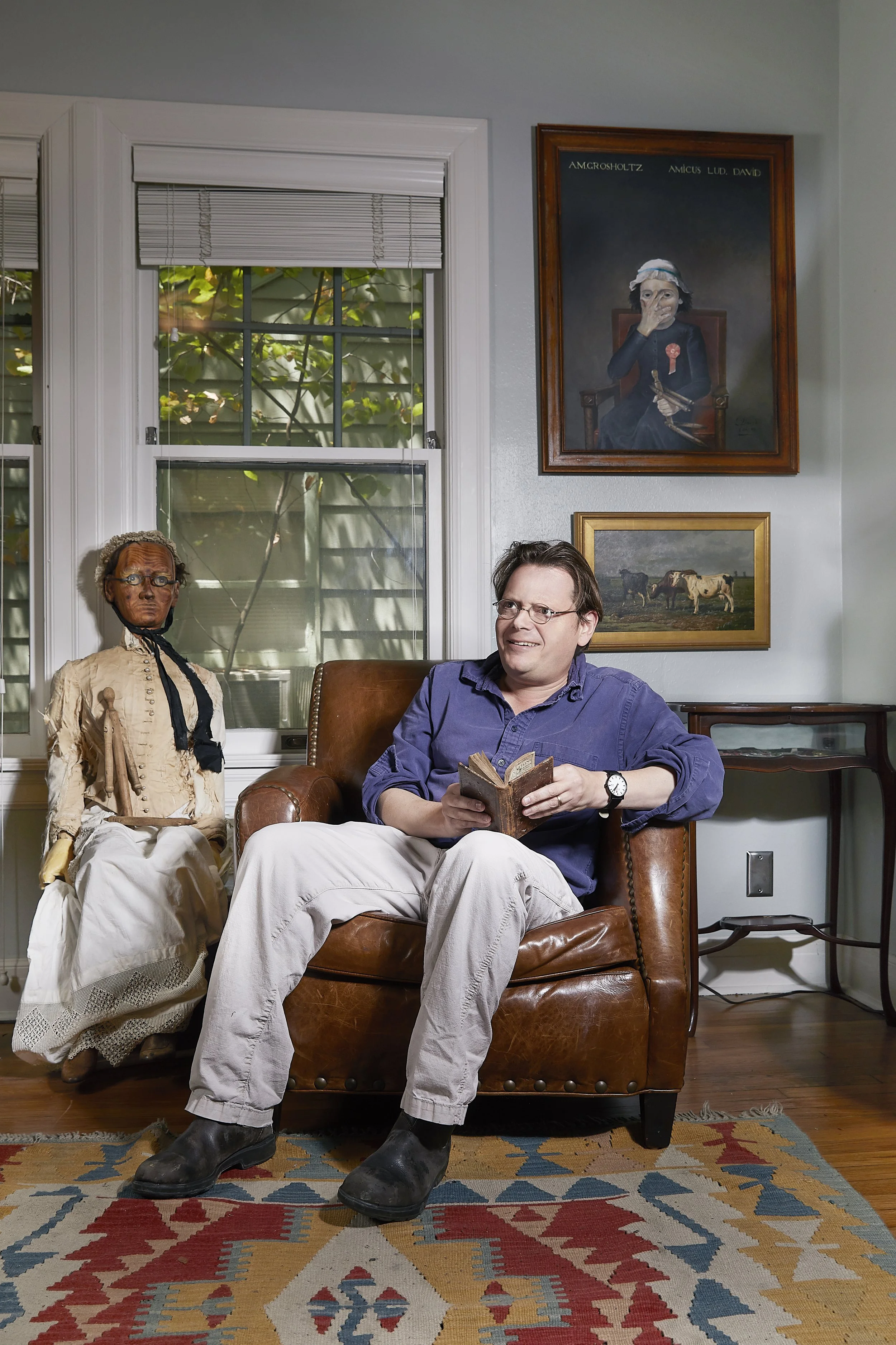 A man sitting in a leather chair holding a book in a living room. A mannequin dressed in old-fashioned clothing is beside him. There is a window with blinds behind them and framed pictures on the wall, including a portrait of a woman and a landscape 
